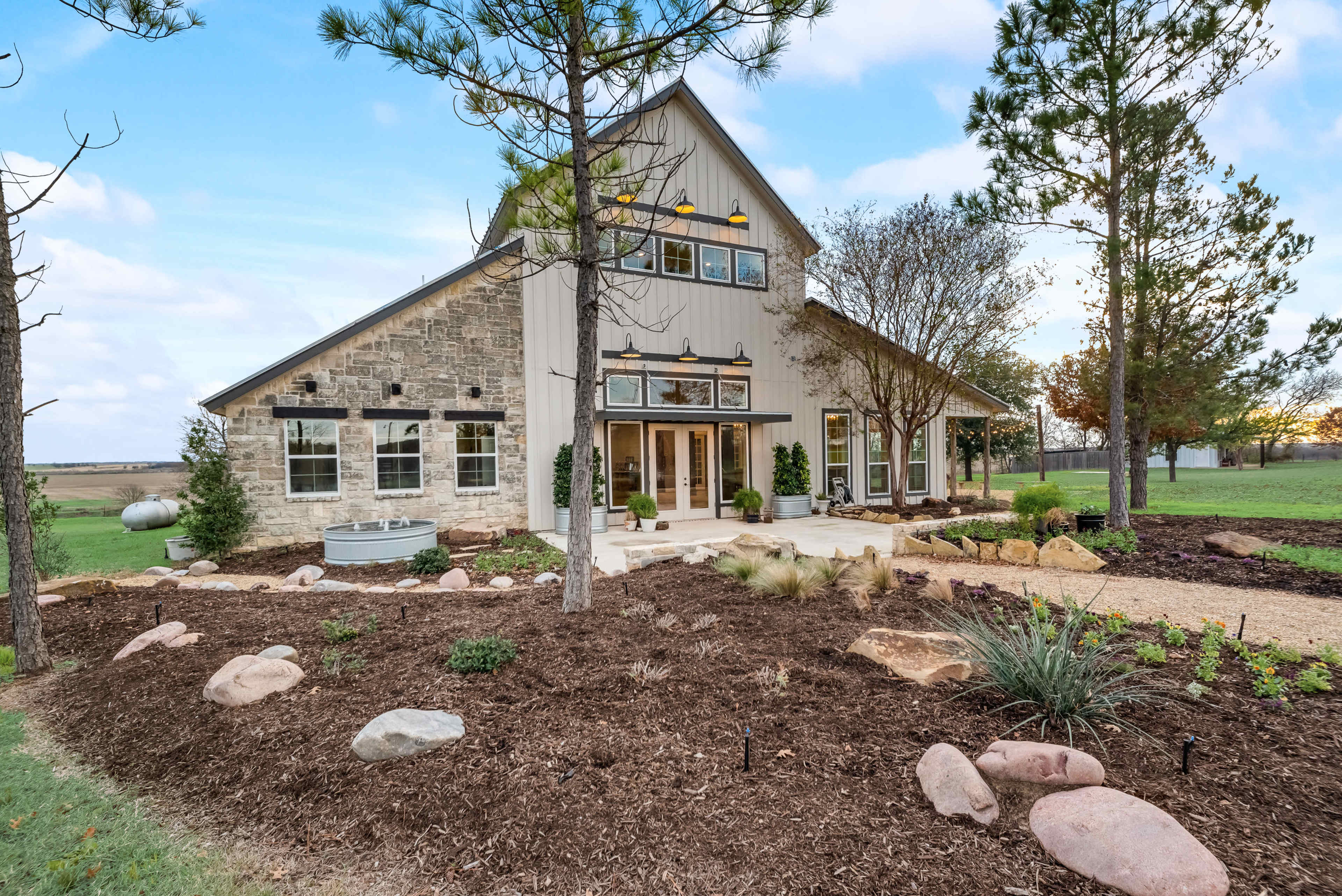 A modern house with a combination of stone and siding exterior sits amidst landscaped gardens featuring rocks, shrubs, and trees.