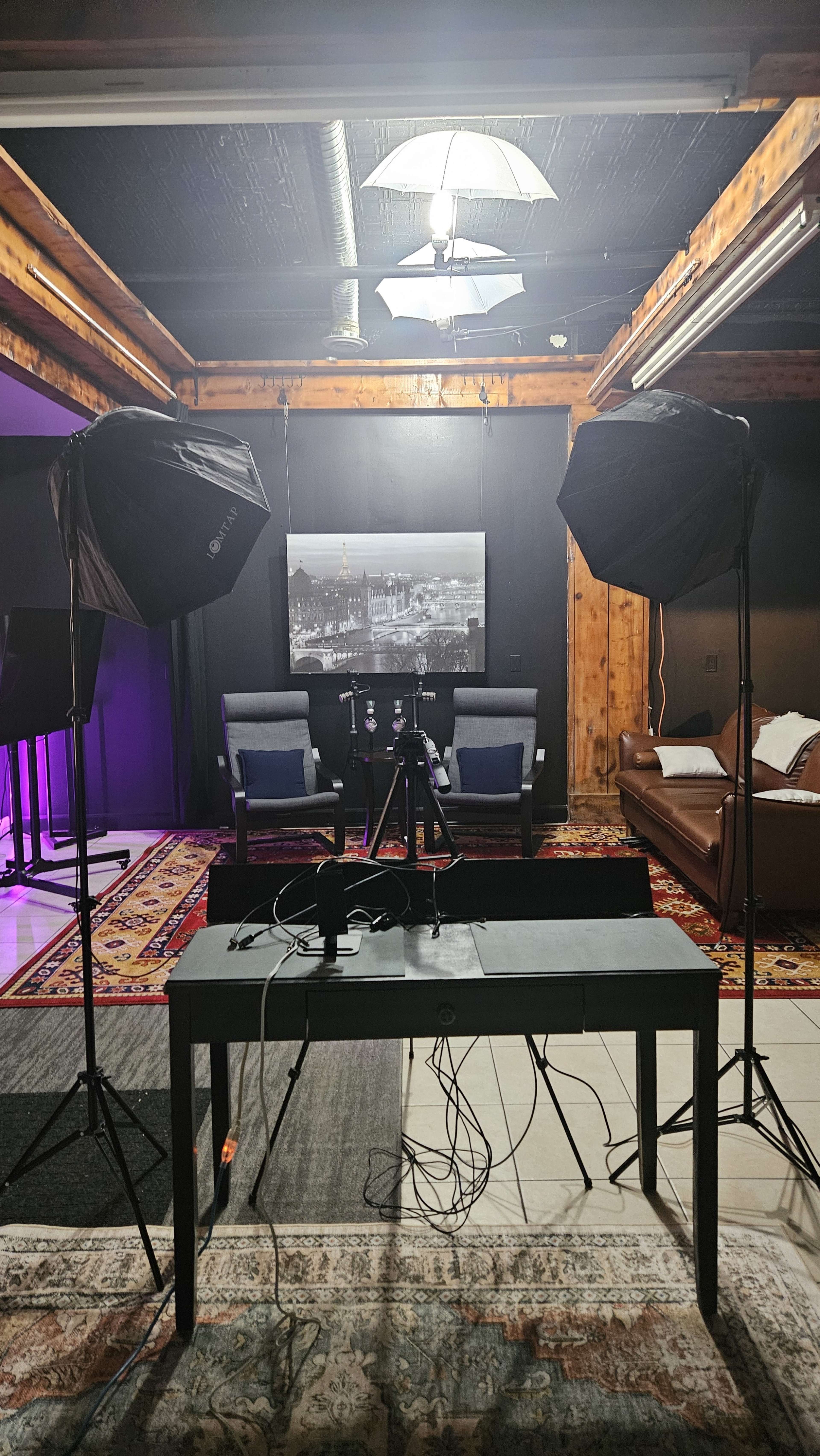 The image shows a recording studio setup featuring two softbox lights, a table with a microphone and camera tripod, and a backdrop of chairs and a framed picture on a wooden wall.