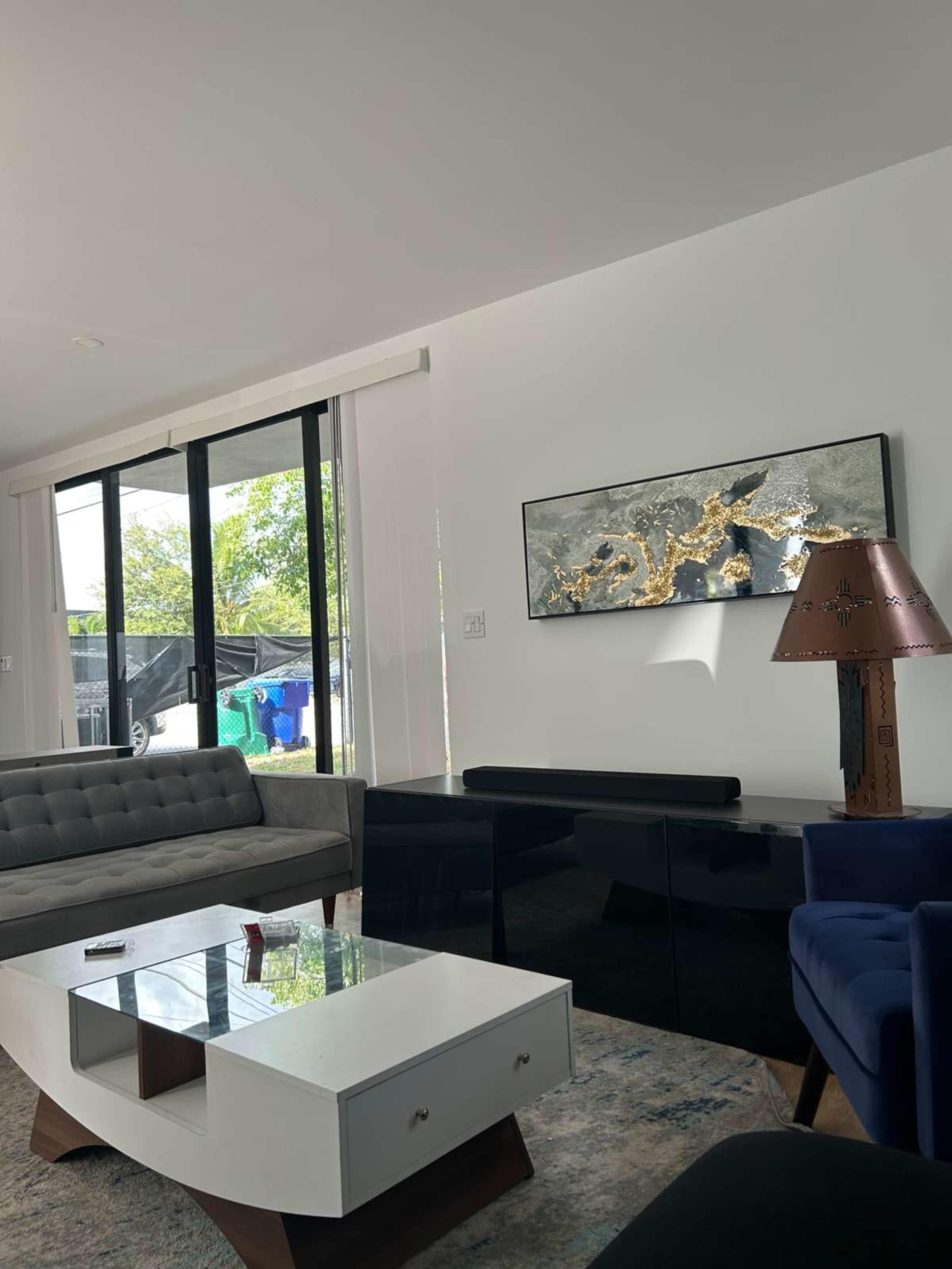 The image shows a modern living room featuring a gray sofa, a white coffee table, a dark glass console, and a decorative lamp beside a large window.