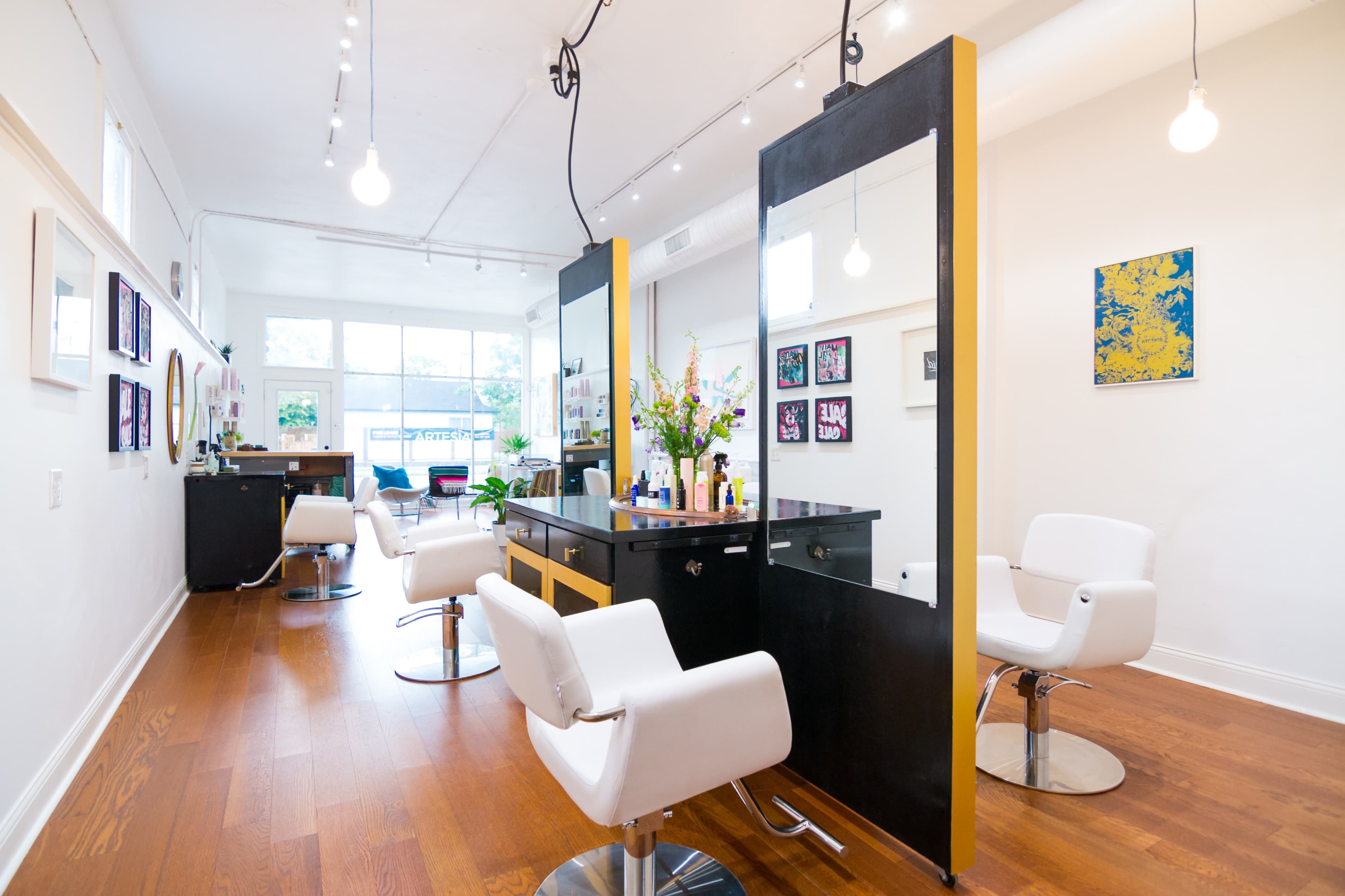 A modern hair salon with two styling stations, equipped with mirrors and white chairs, set against a backdrop of light wood flooring and bright lighting.