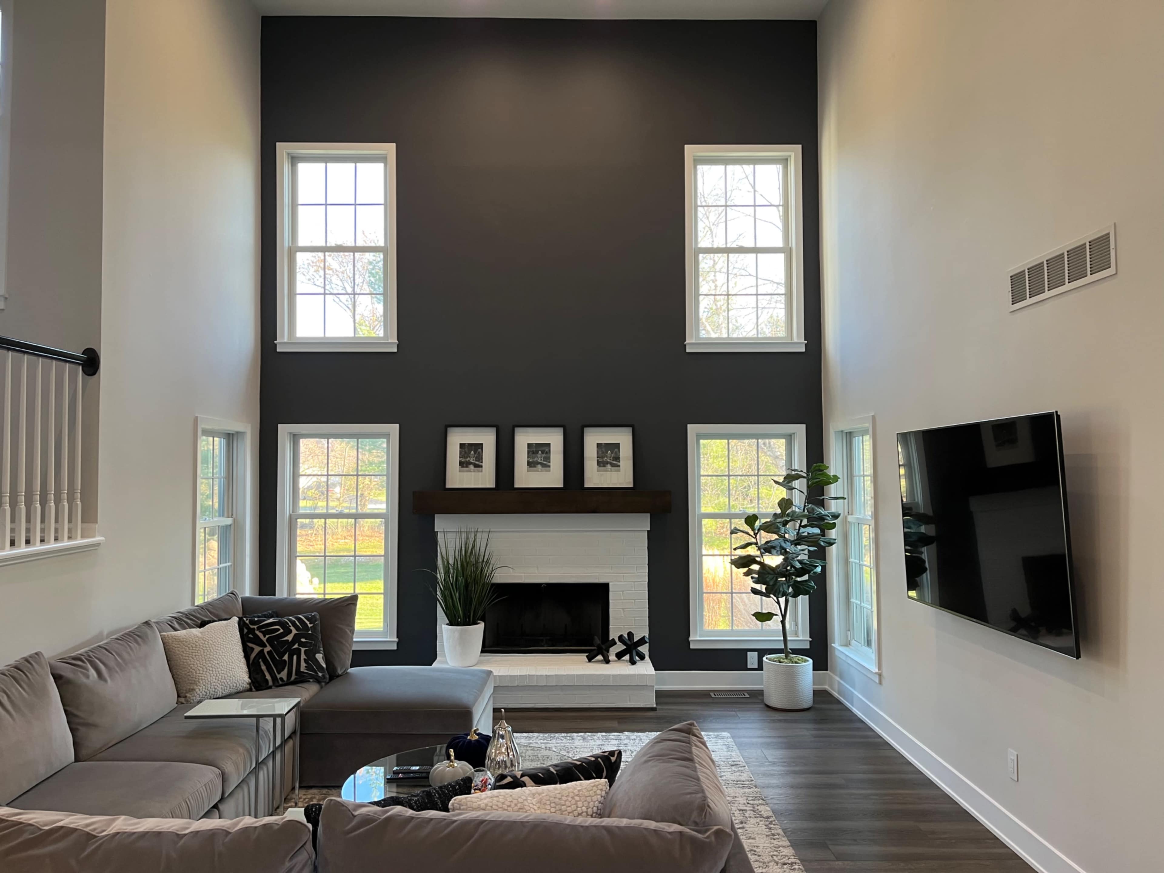 The image shows a modern living room with a tall gray accent wall, large windows, a white fireplace, a sectional sofa, and a television mounted on the wall.