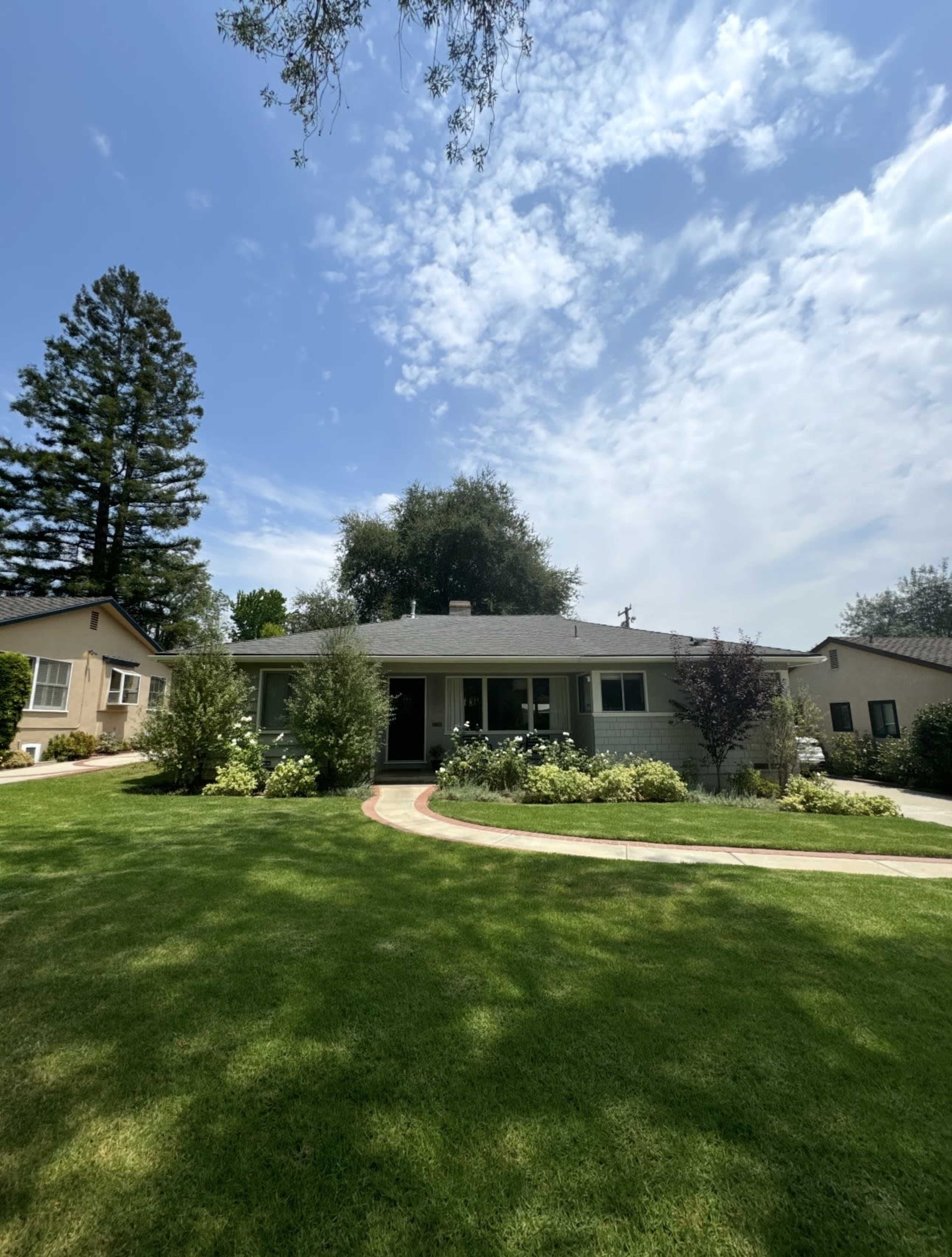 A single-story home with a landscaped yard and a curved pathway leads to the entrance, surrounded by trees and shrubs under a partly cloudy sky.
