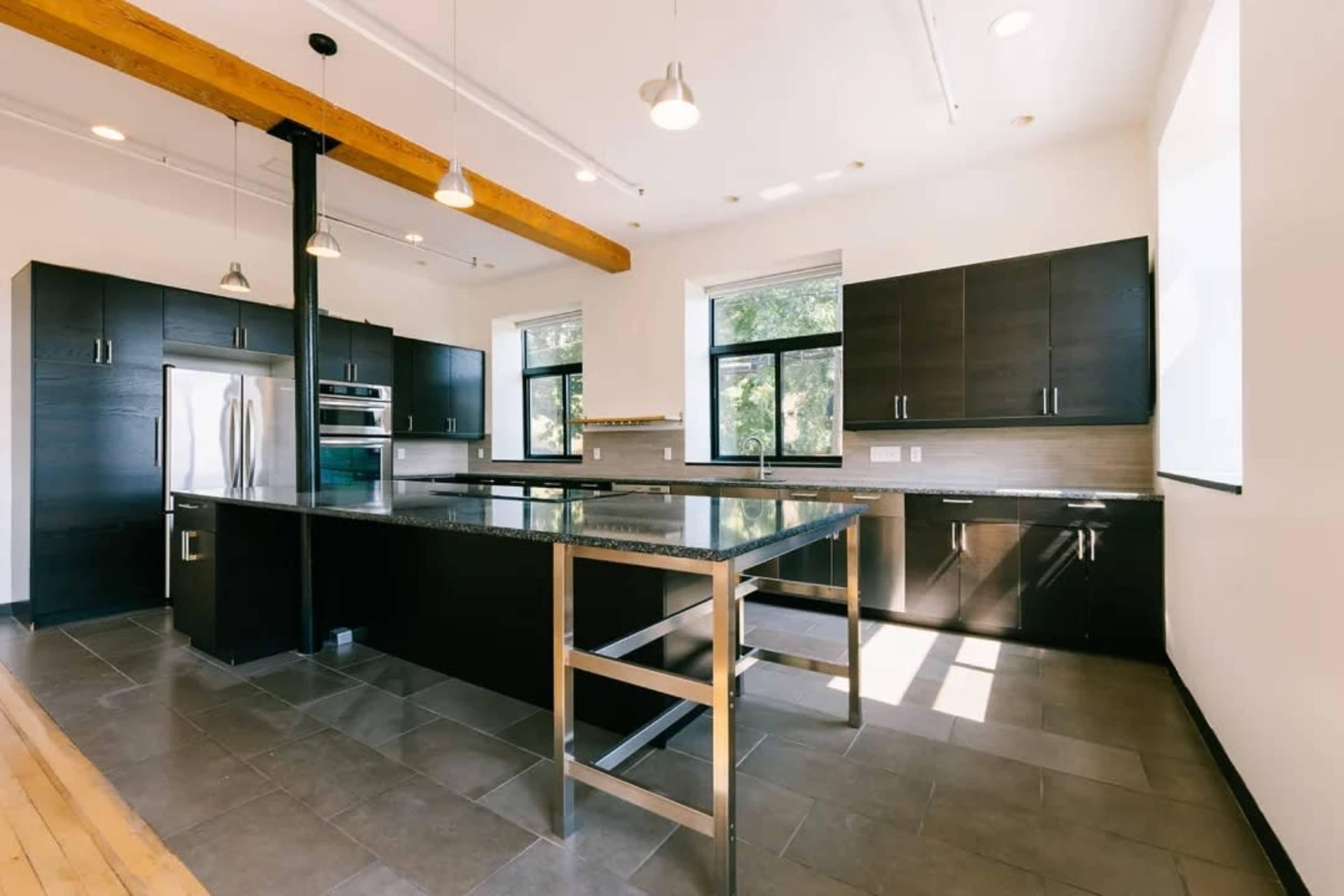 The image shows a modern kitchen featuring black cabinets, a large granite countertop island, and stainless steel appliances, with large windows allowing natural light.