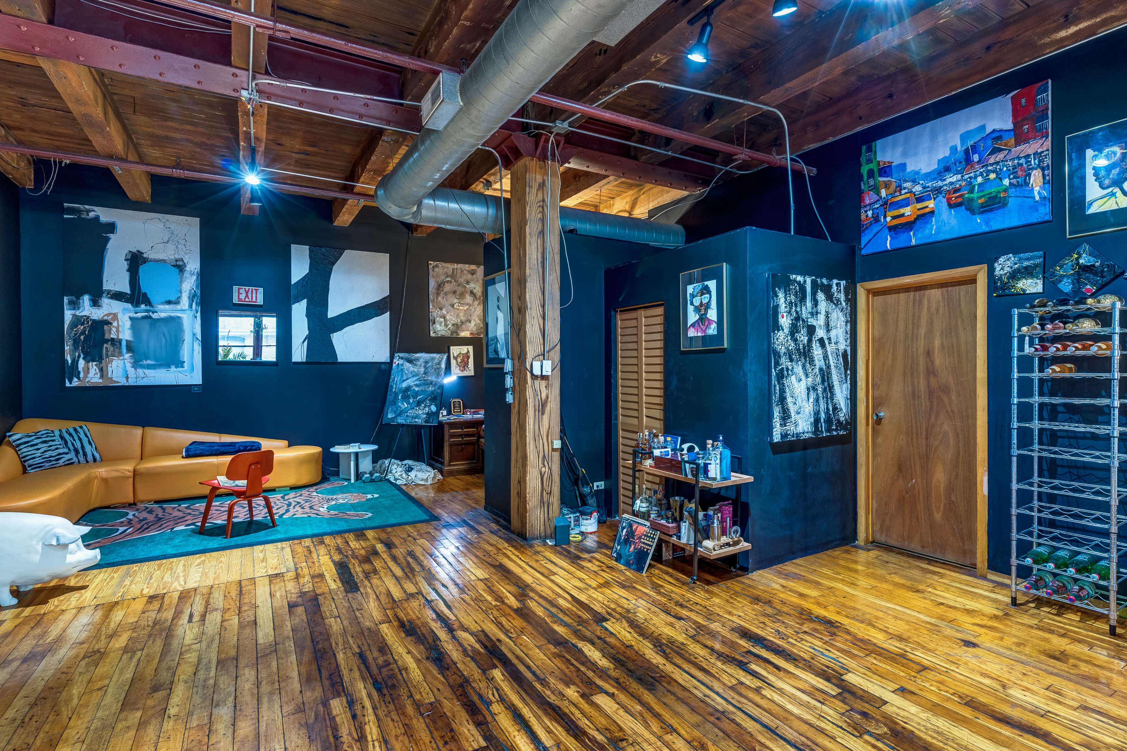 The scene shows a spacious room with wooden floors, exposed beams, and black walls adorned with various artworks, featuring a couch, a small red chair, and shelves displaying art supplies.