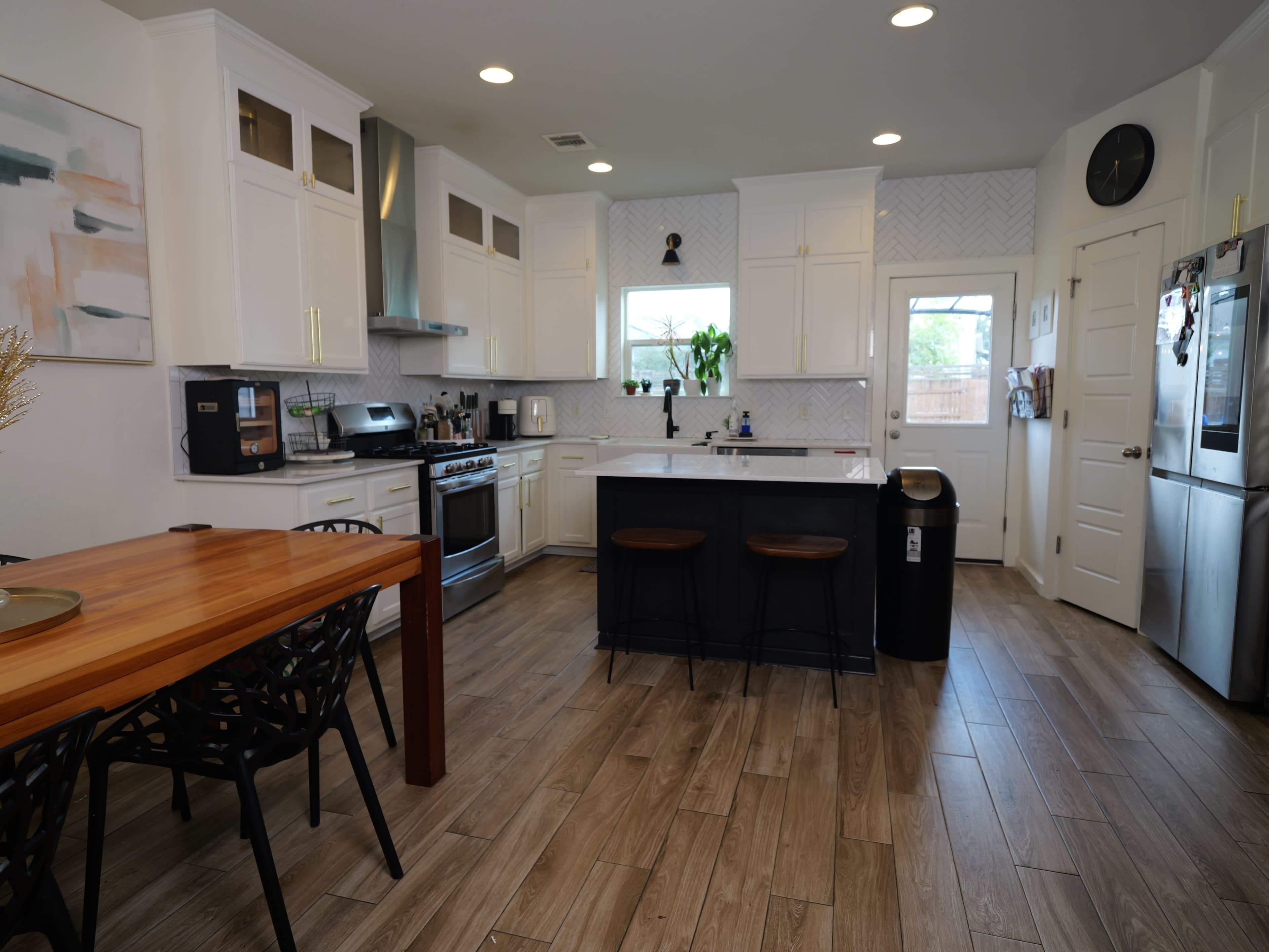 The image shows a modern kitchen with white cabinets, a large island with bar stools, and a dining table beside it.
