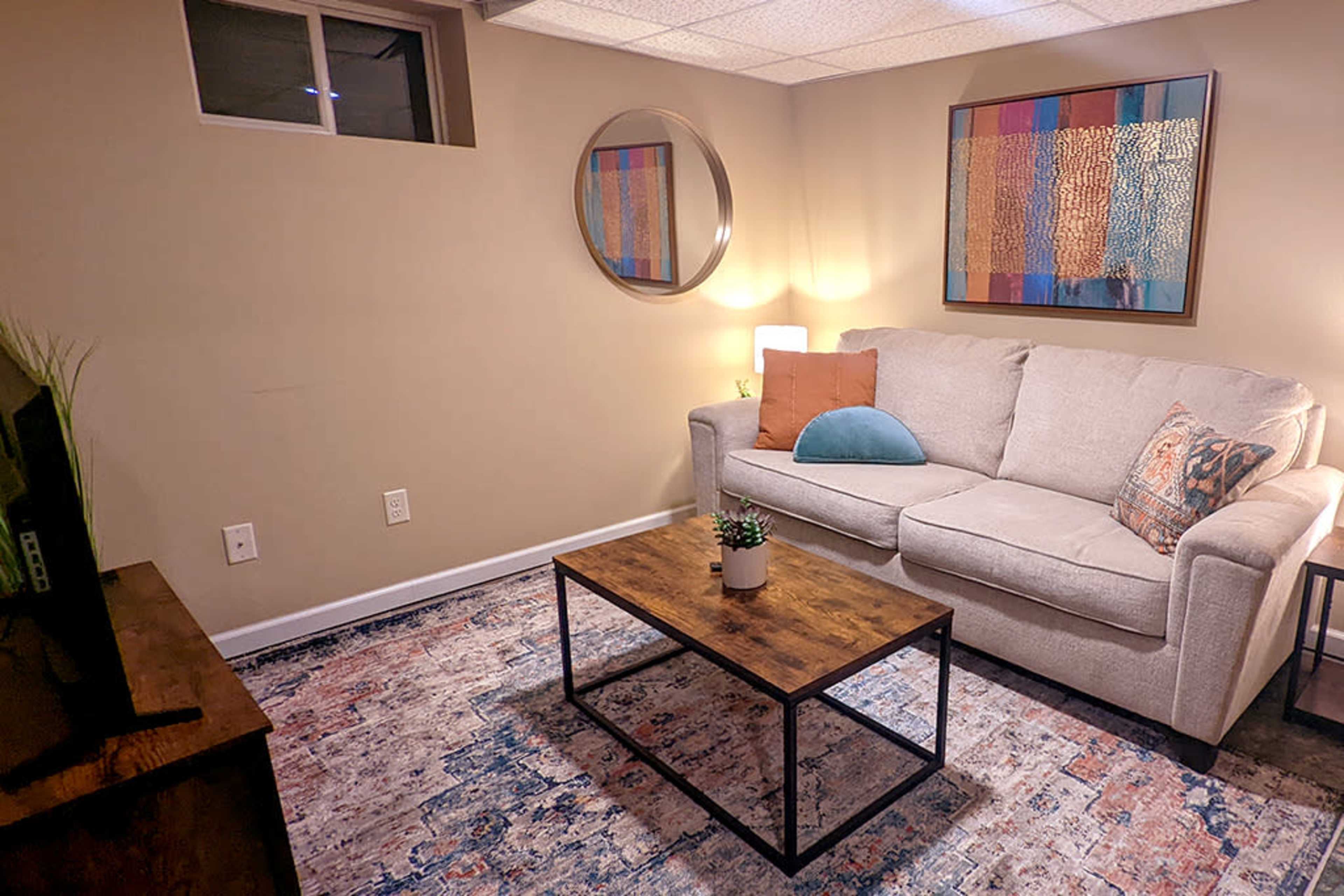 The image shows a small, neatly arranged living room with a light gray sofa, a wooden coffee table, a decorative mirror, and a colorful area rug.