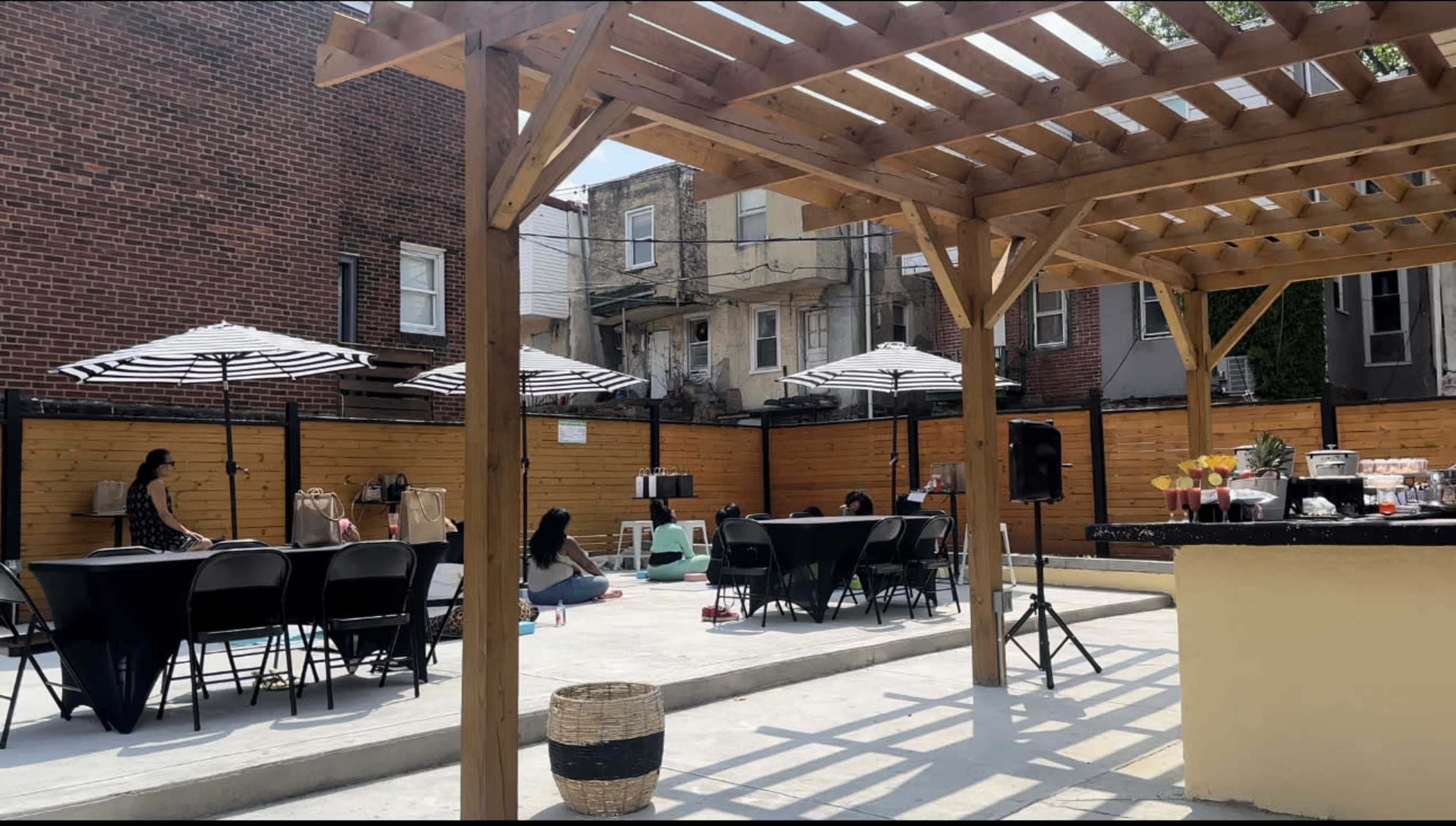 The scene shows an outdoor patio area with black tables and chairs, striped umbrellas, and a wooden pergola, while several individuals are seated in the background.