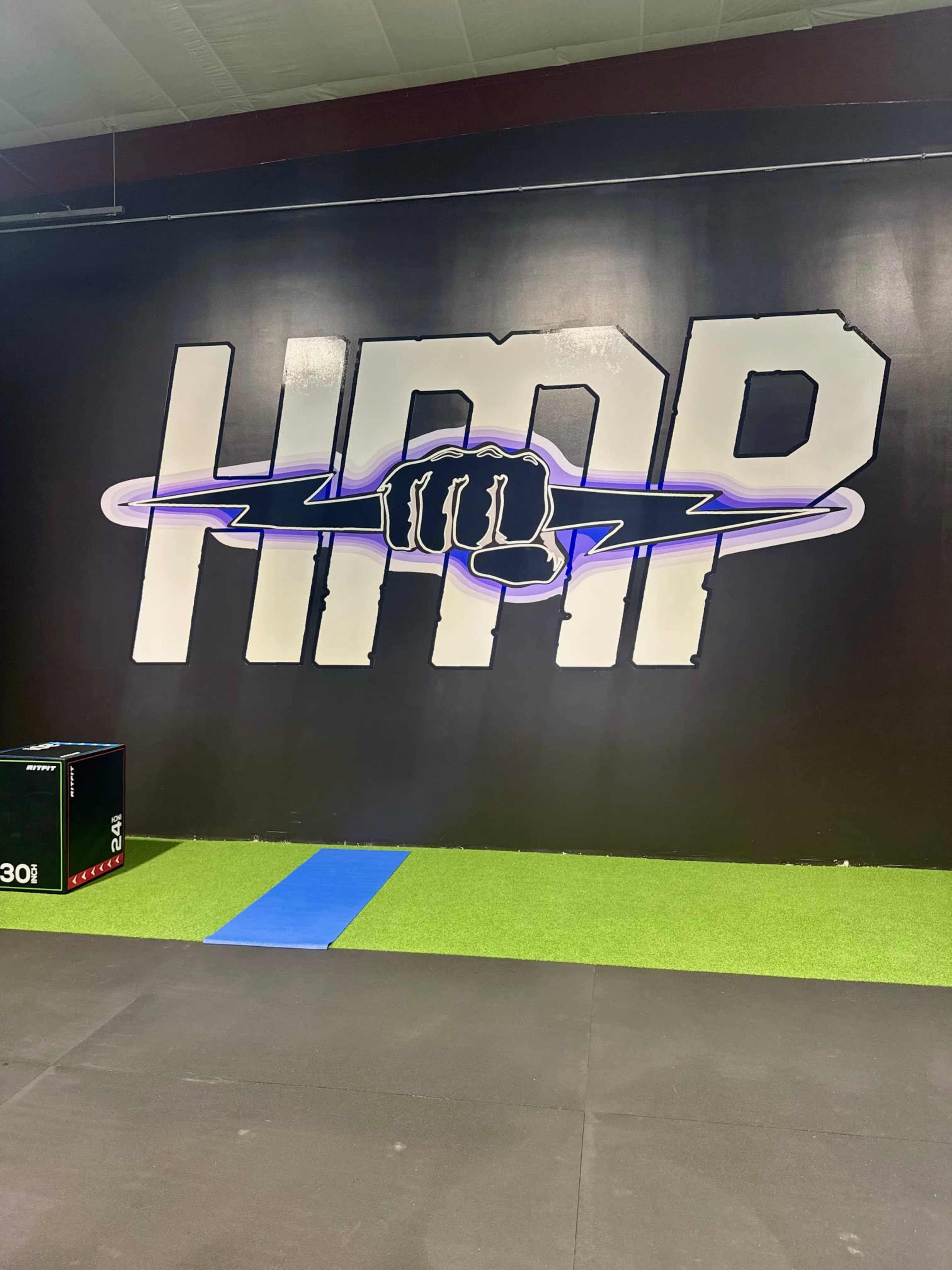 The image shows a black wall with a large, stylized logo featuring a clenched fist and the letters "HIMP," next to a blue exercise mat and a green turf section on the floor.