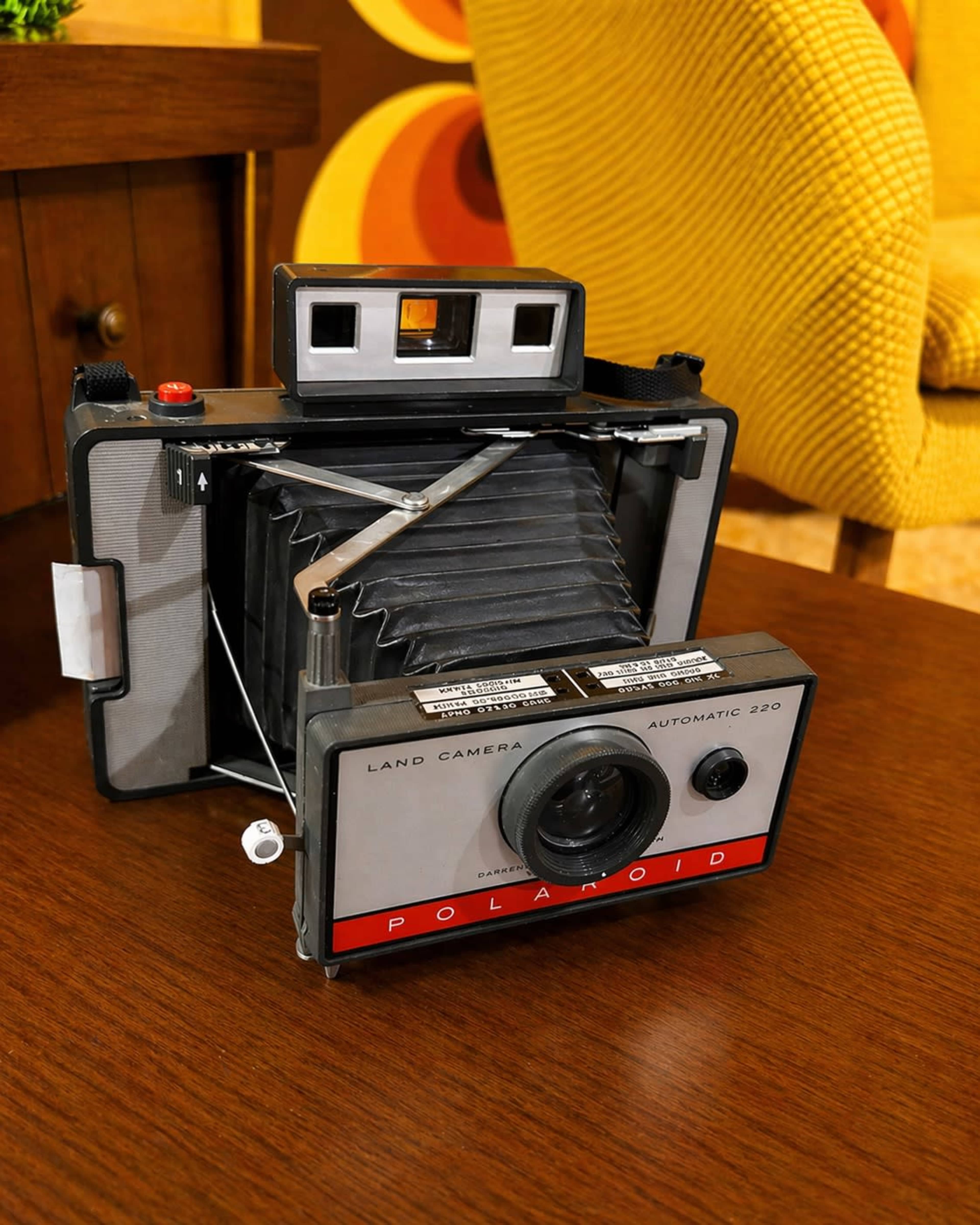 The image shows an old Polaroid Land Camera Automatic 220 positioned on a wooden table with a textured background.