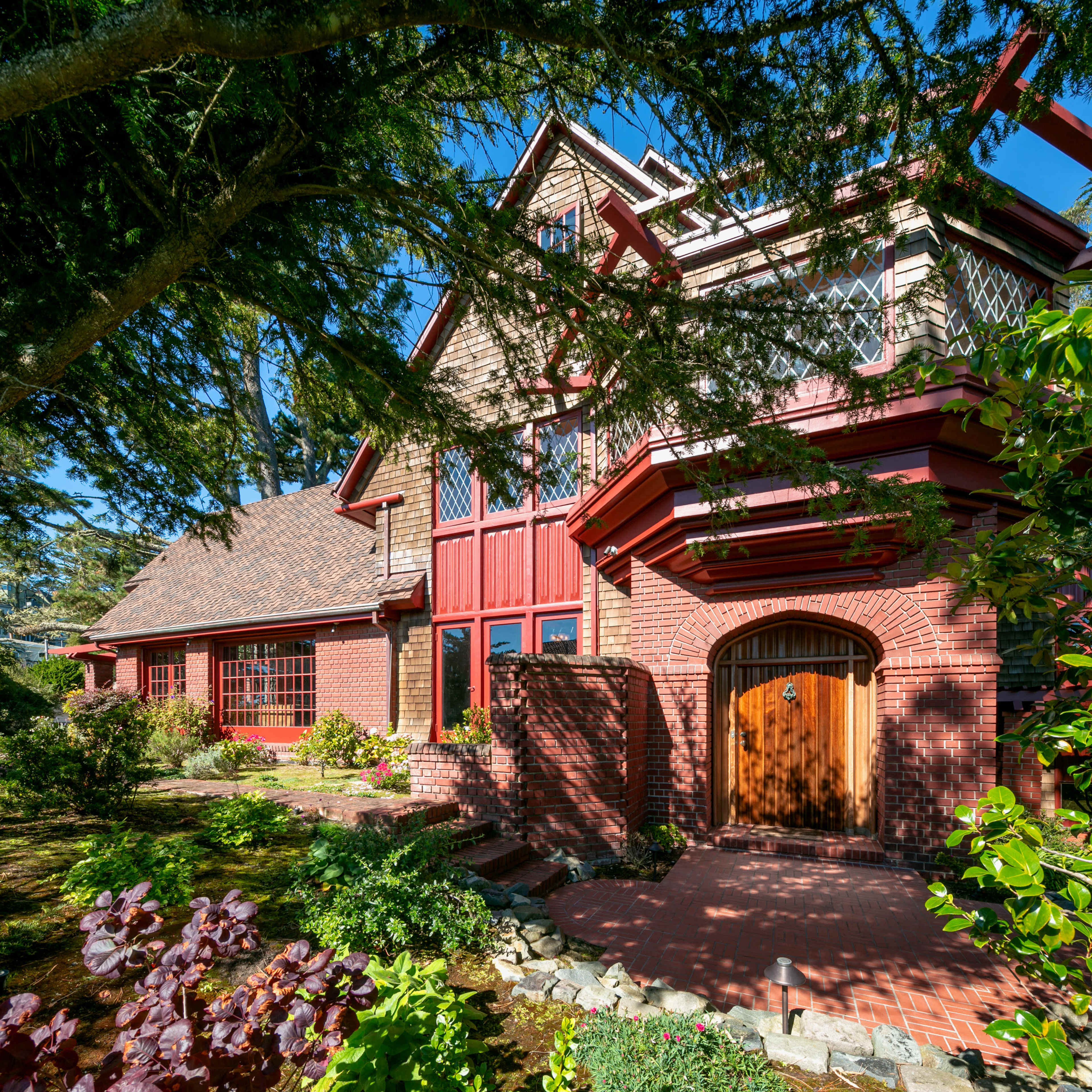 A large, two-story brick house with red accents and a wooden front door, surrounded by lush greenery and a stone pathway.