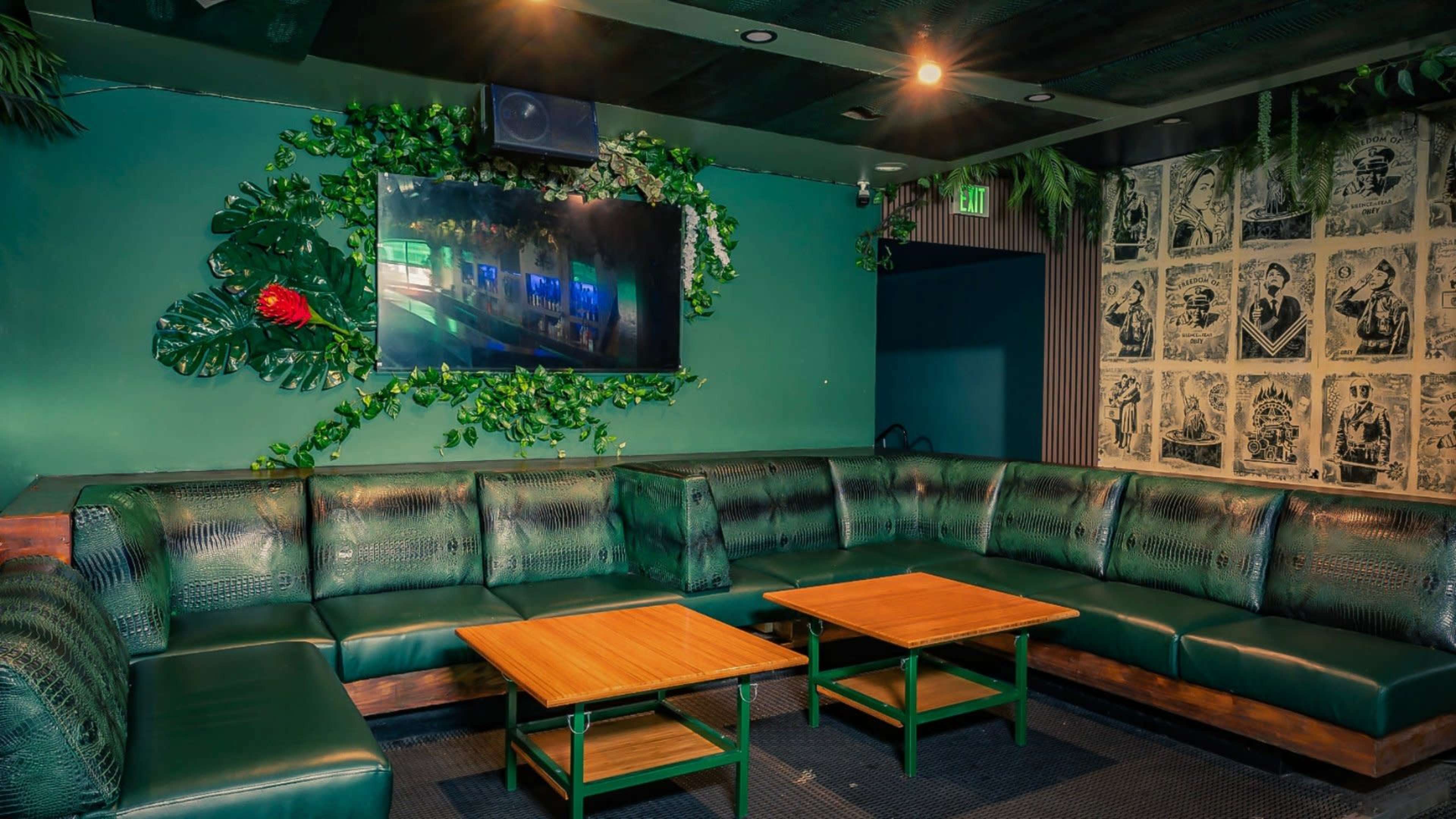 The image shows a spacious lounge area with green leather seating, two wooden tables, and a wall decorated with plants and a television.