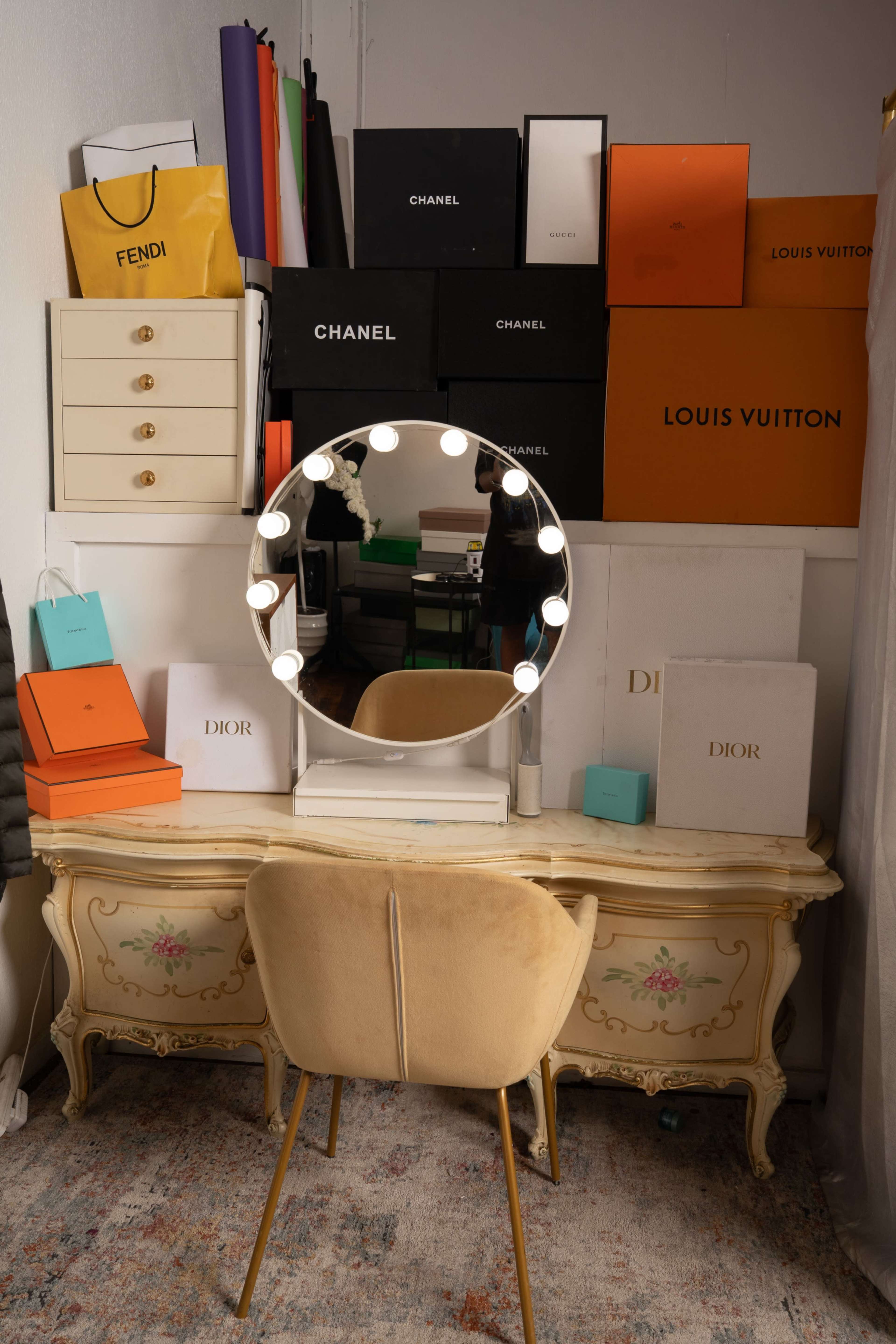 A vintage vanity with a round mirror and bright light bulbs is positioned against a wall adorned with various designer brand boxes.