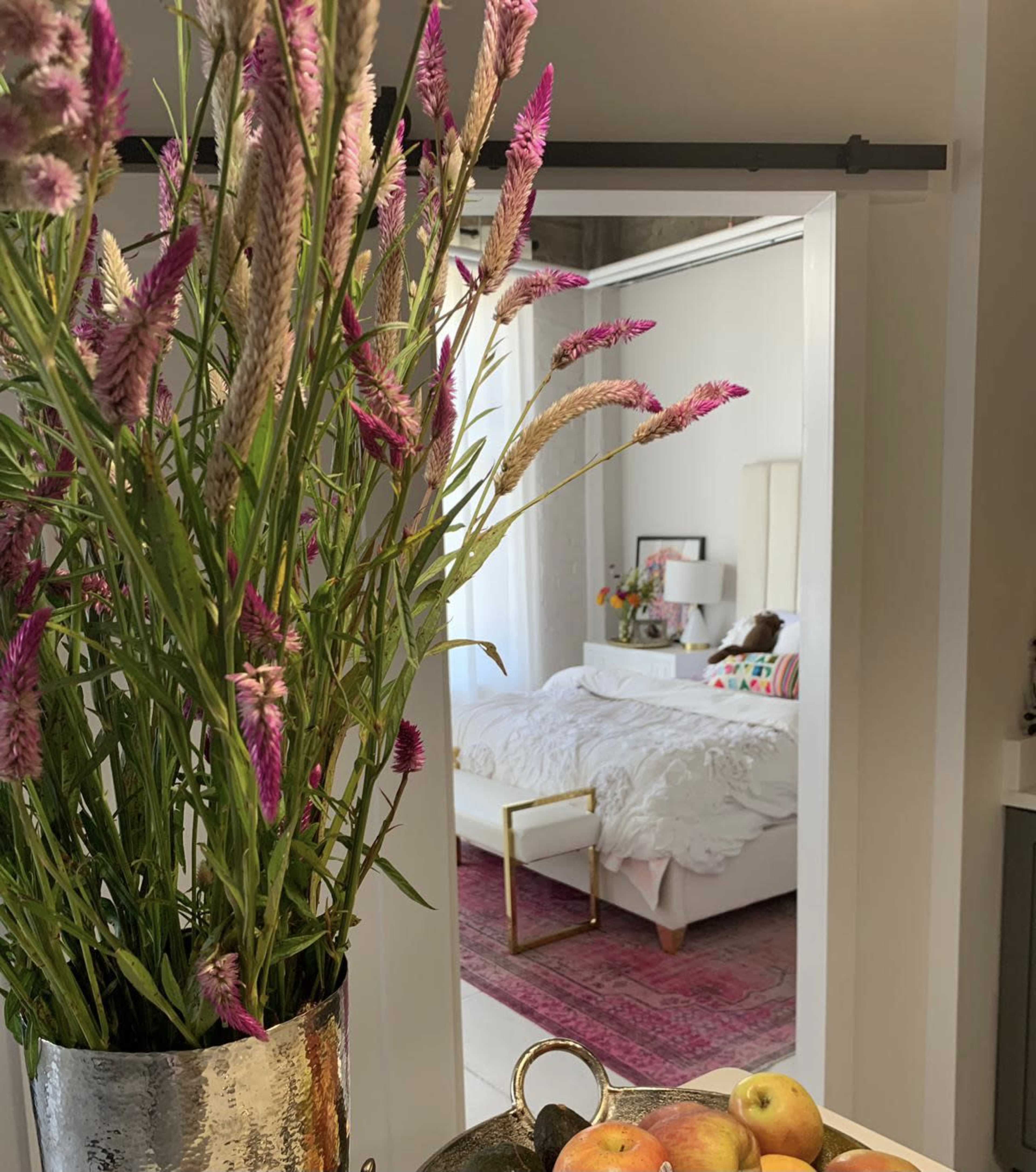 A bouquet of pink flowers in a vase is placed in the foreground, with a glimpse of a bedroom featuring a bed and colorful decor in the background.