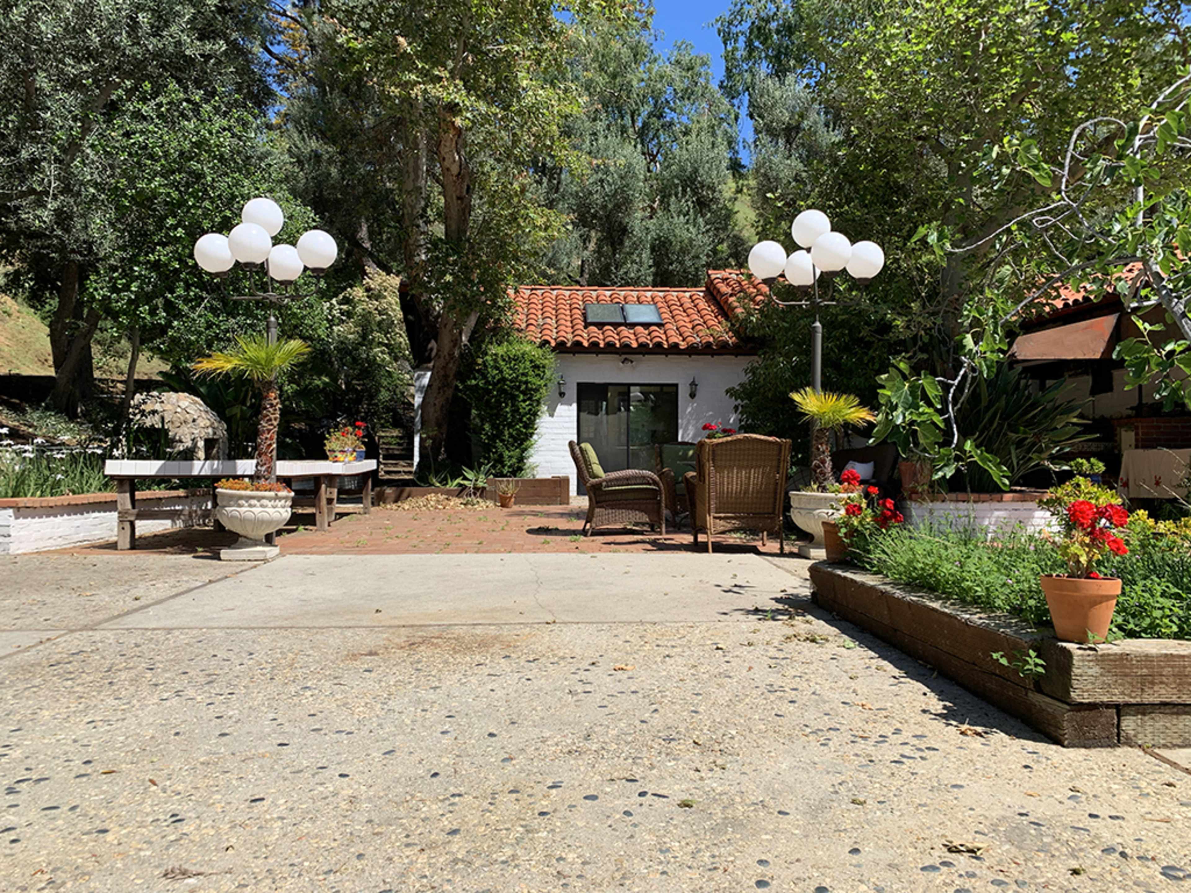 The image shows a spacious outdoor area featuring a mix of seating arrangements, decorative plants, and a house with a red tile roof, surrounded by trees.