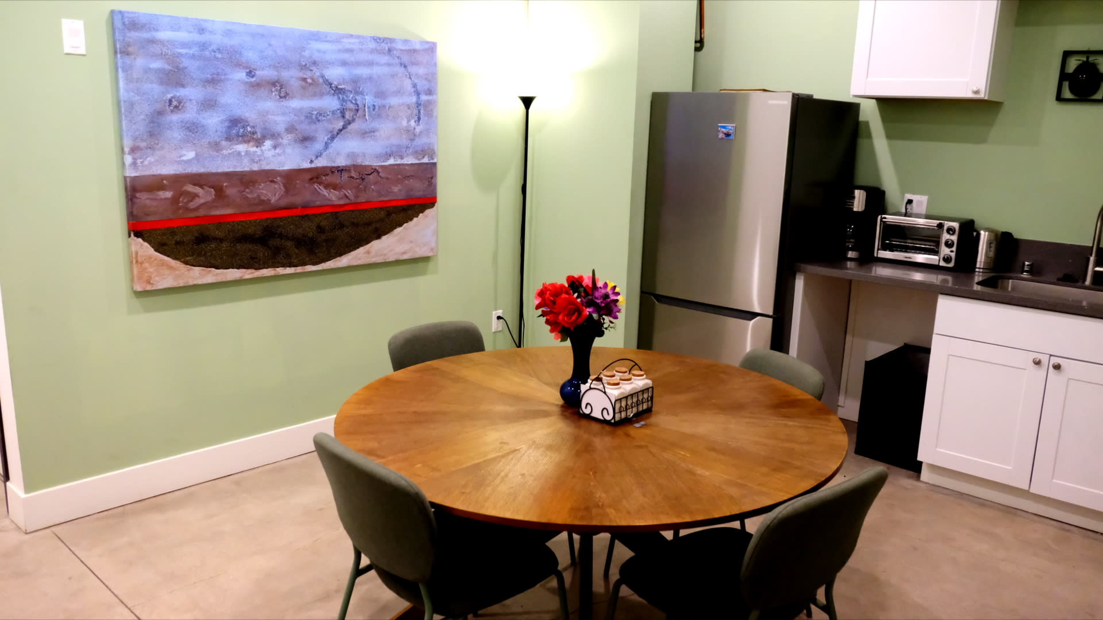 The image shows a kitchen area with a round wooden dining table and four green chairs, a refrigerator, and an abstract painting on the wall.