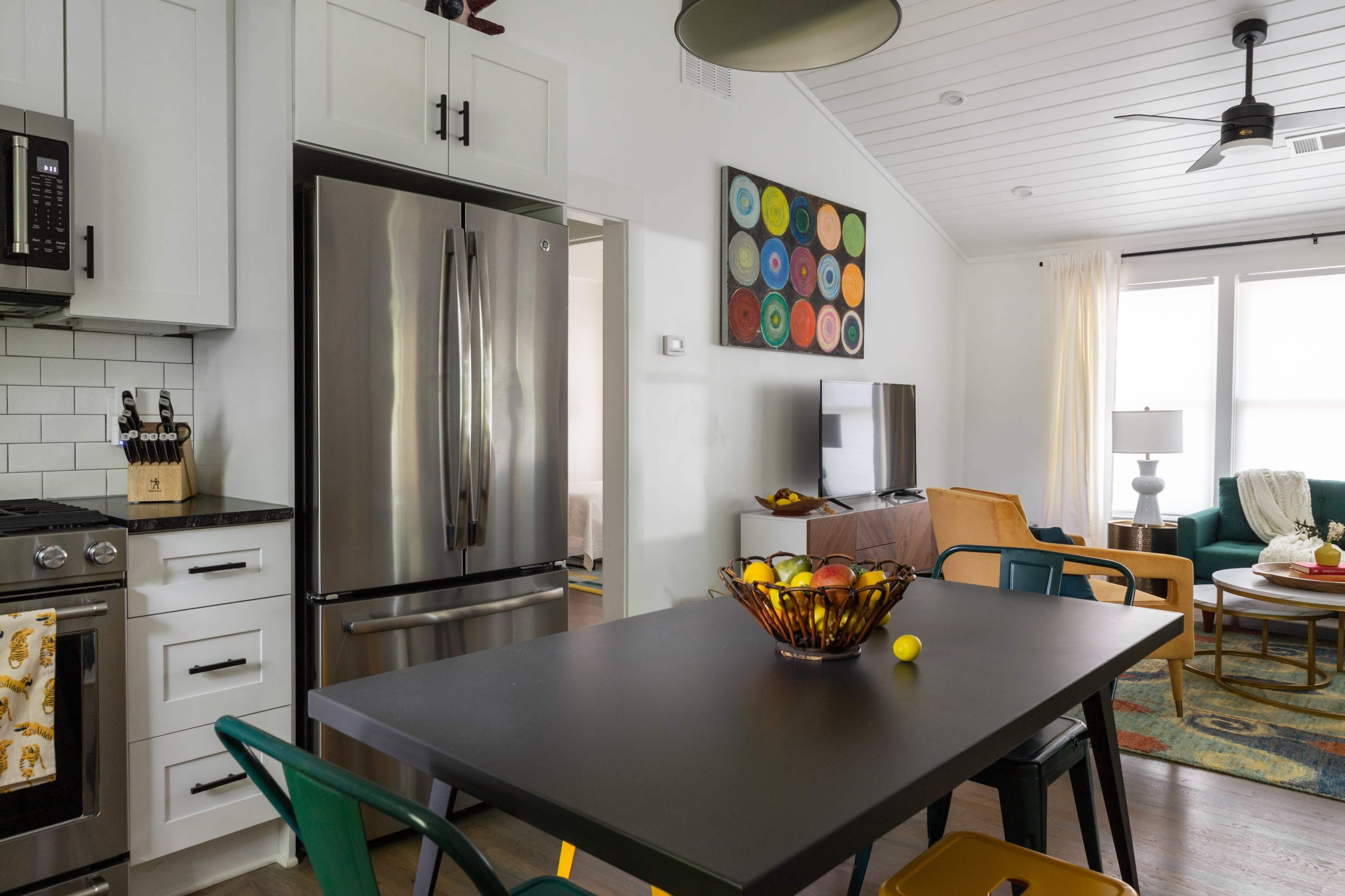 A modern kitchen and living area features a black dining table with a bowl of fruit, stainless steel appliances, and a colorful wall art above, along with a cozy seating area.