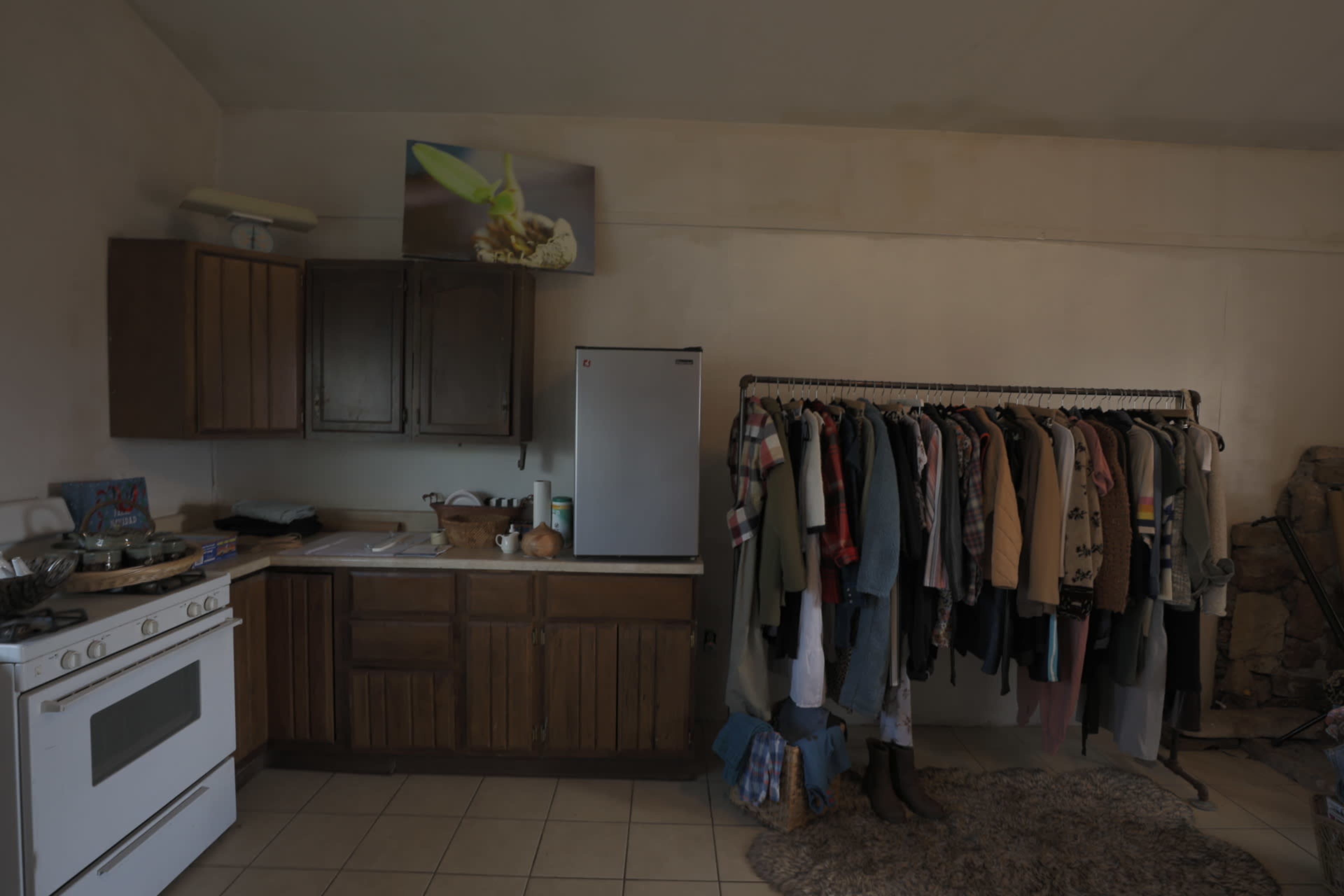 The image shows a kitchen area with wooden cabinets, a refrigerator, and a clothing rack filled with various garments.