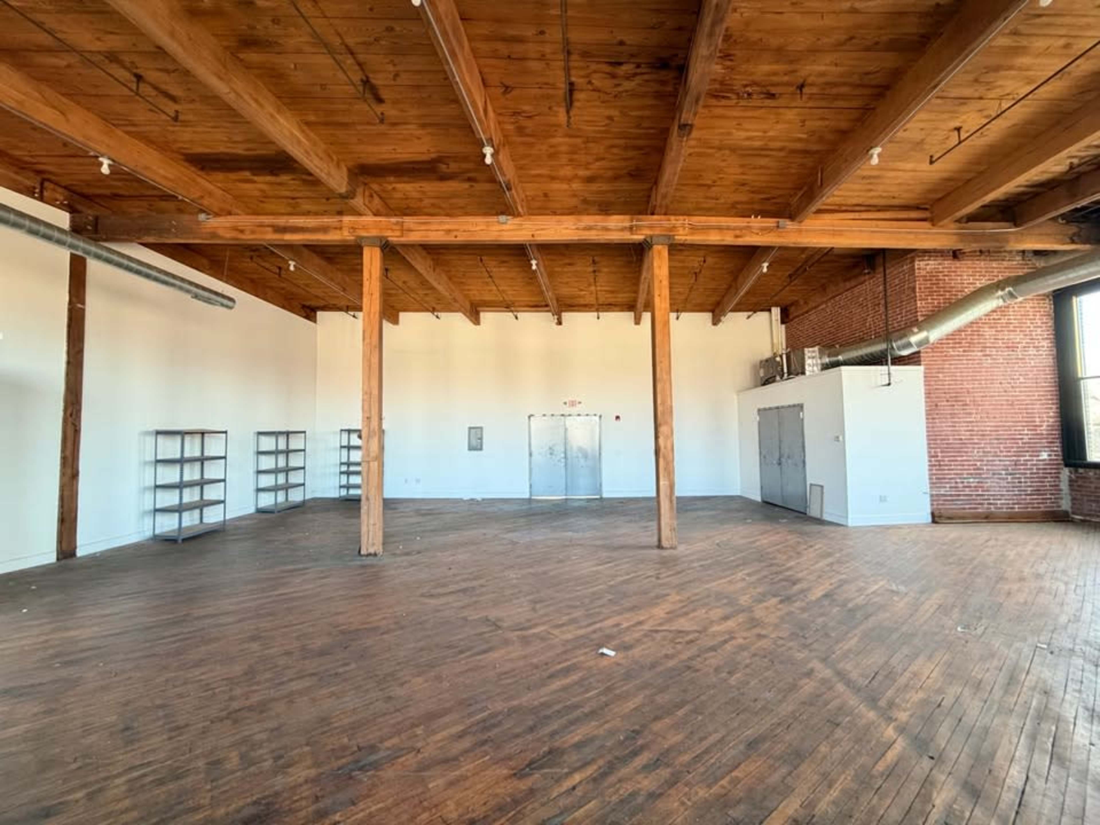 The image shows an empty industrial-style room with wooden beams, a wooden floor, and large windows, featuring a few shelves along one wall.