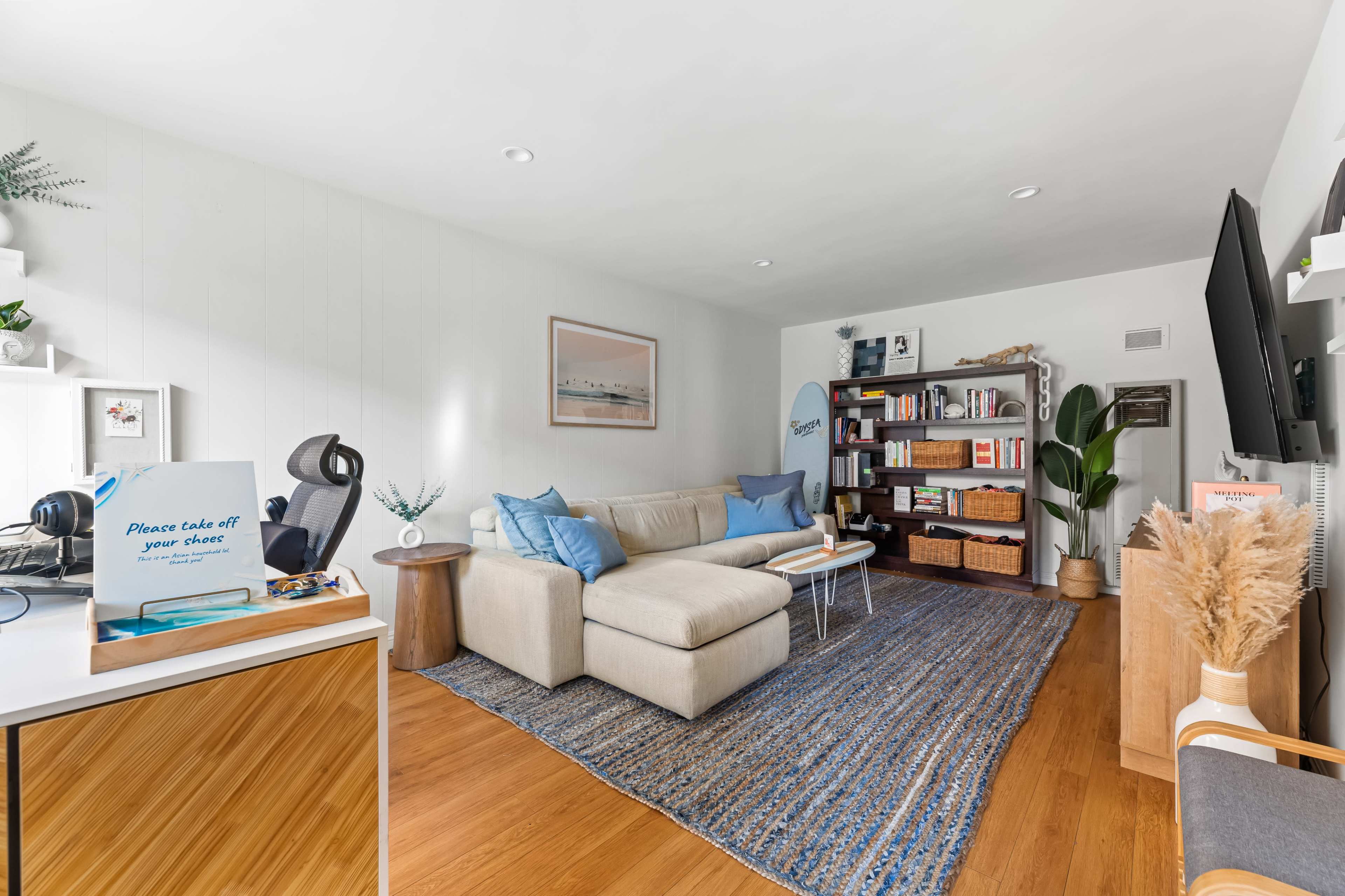 The image shows a cozy living room featuring a light-colored sectional sofa, a wooden bookshelf filled with books and decor, and a flat-screen TV mounted on a wall.