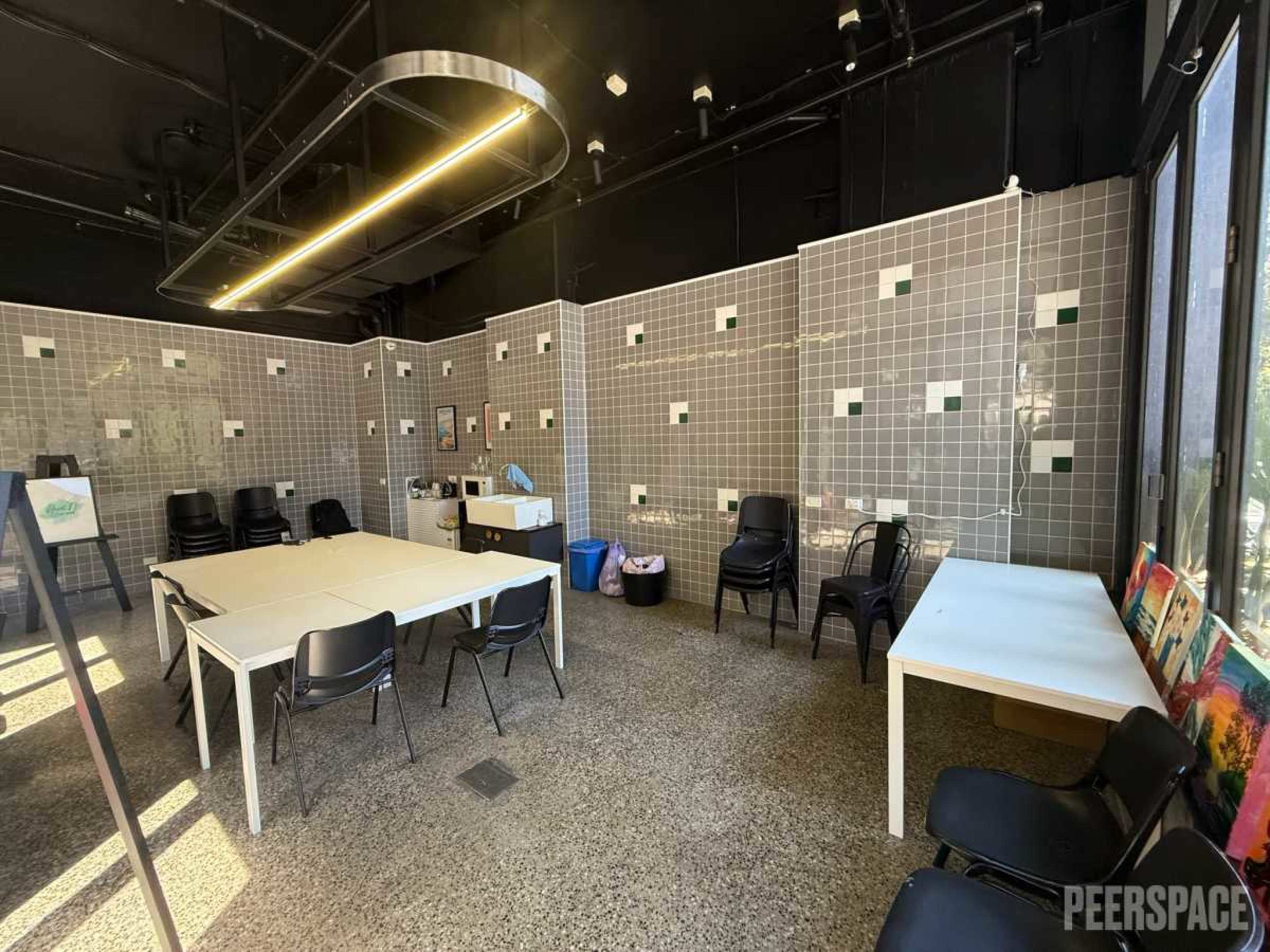 The image shows a modern, spacious room with tiled walls, a large table, several black chairs, and windows allowing natural light to enter.
