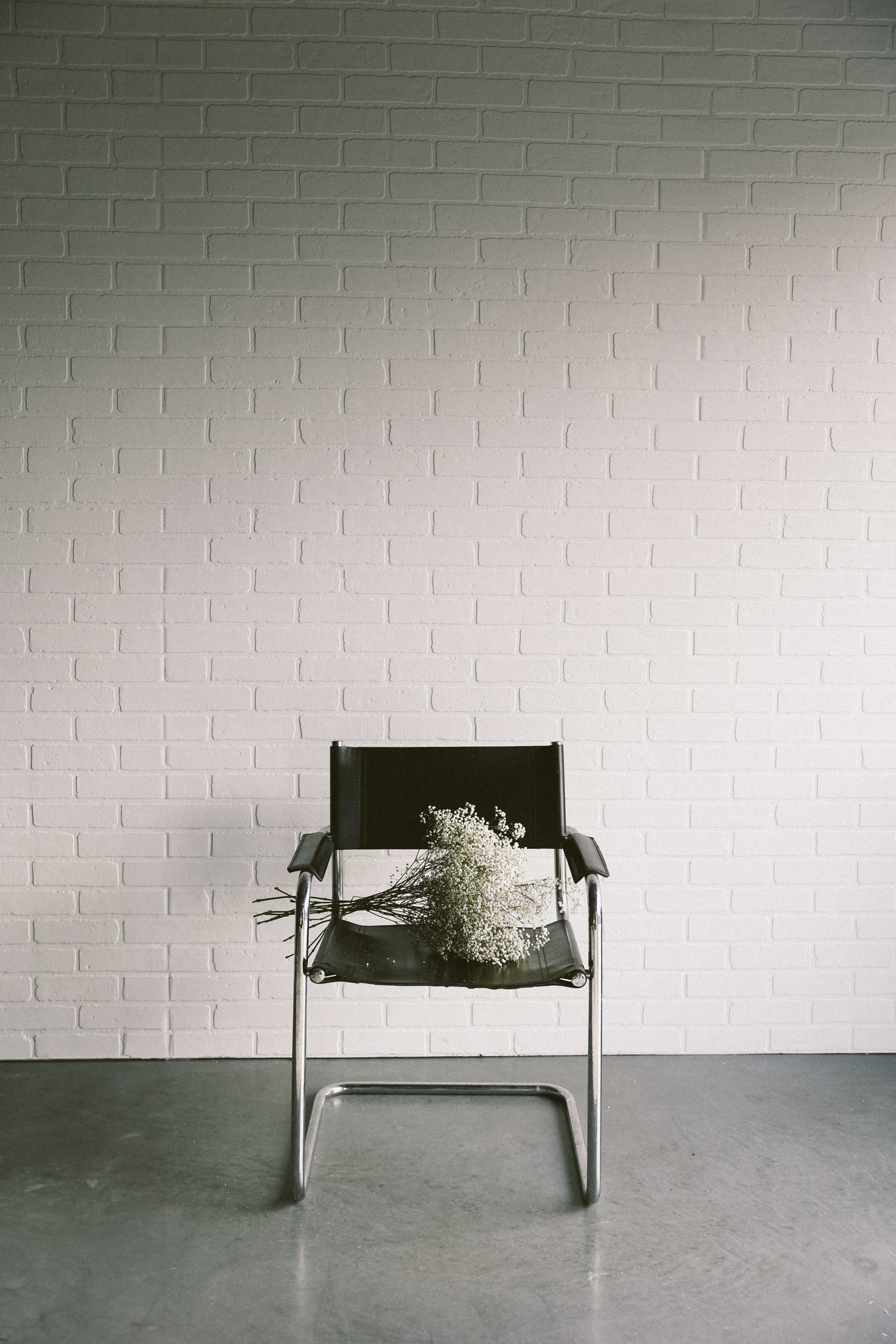 A black chair with metal legs sits against a white brick wall, adorned with a bouquet of dried flowers.