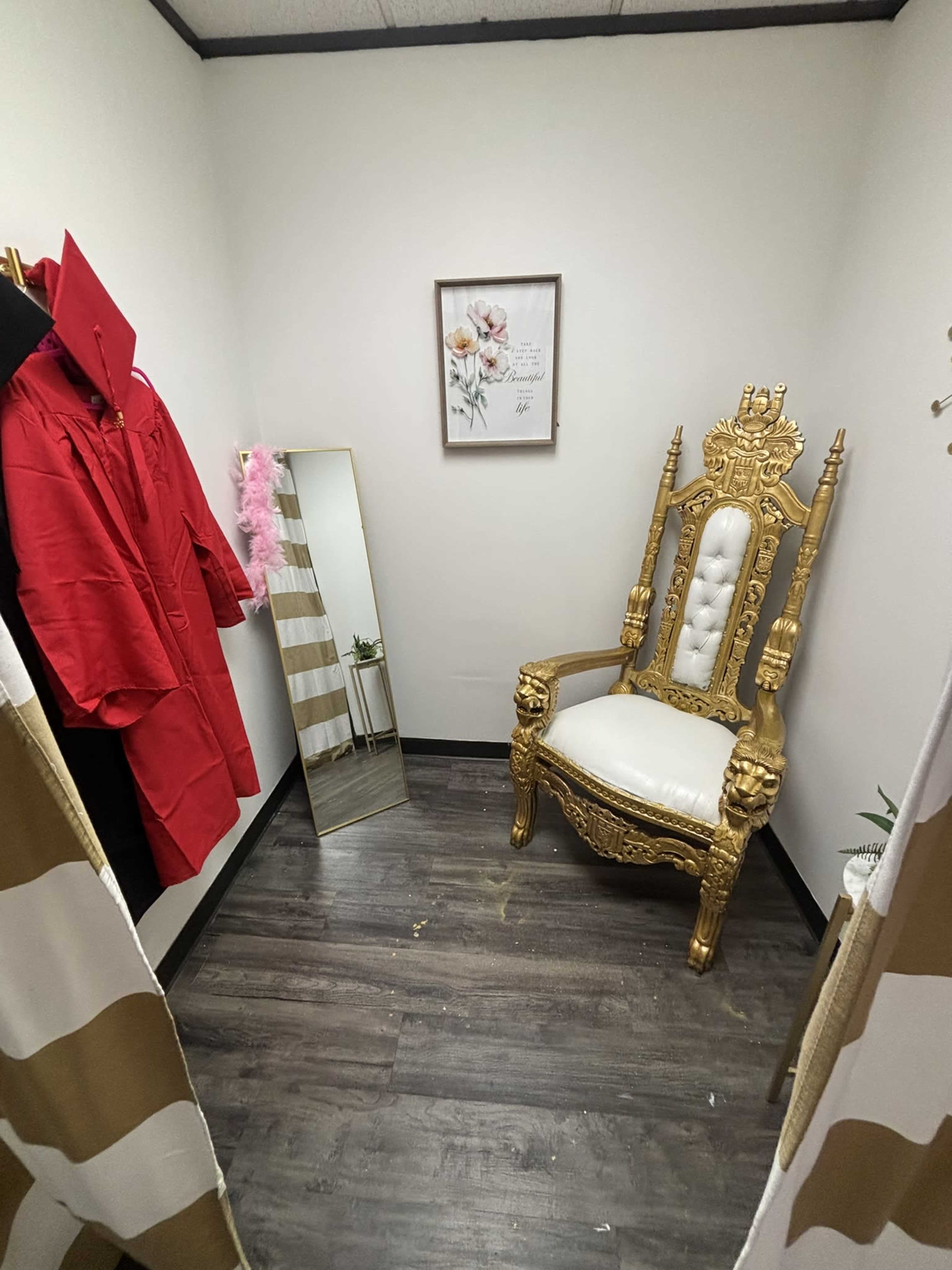 The image shows a small room featuring a gold and white throne chair, a tall mirror, a framed floral artwork, and red robes hanging on the wall.