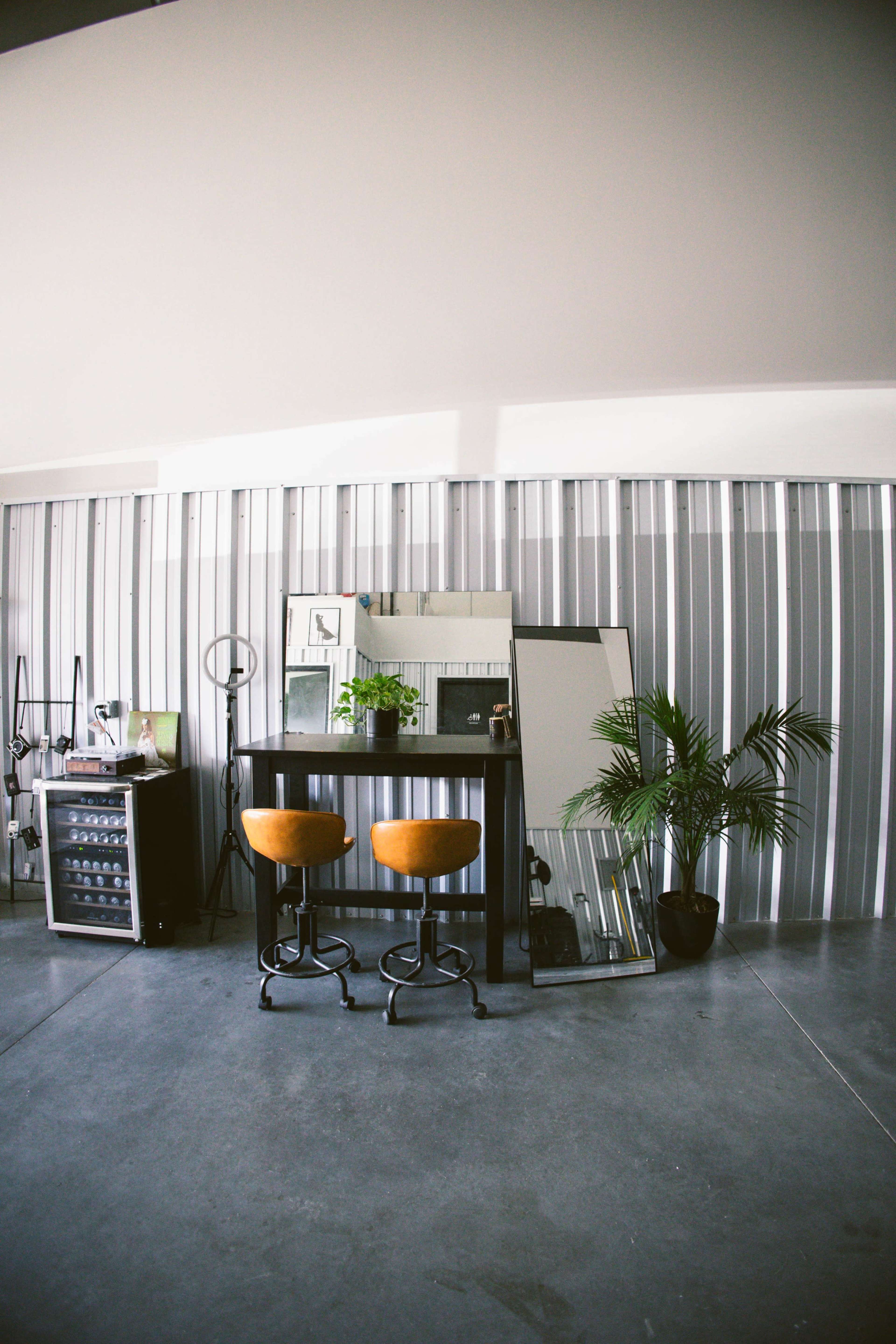 A hair salon features two orange bar stools in front of a large mirrored wall, with a plant and a styling station visible in the background.