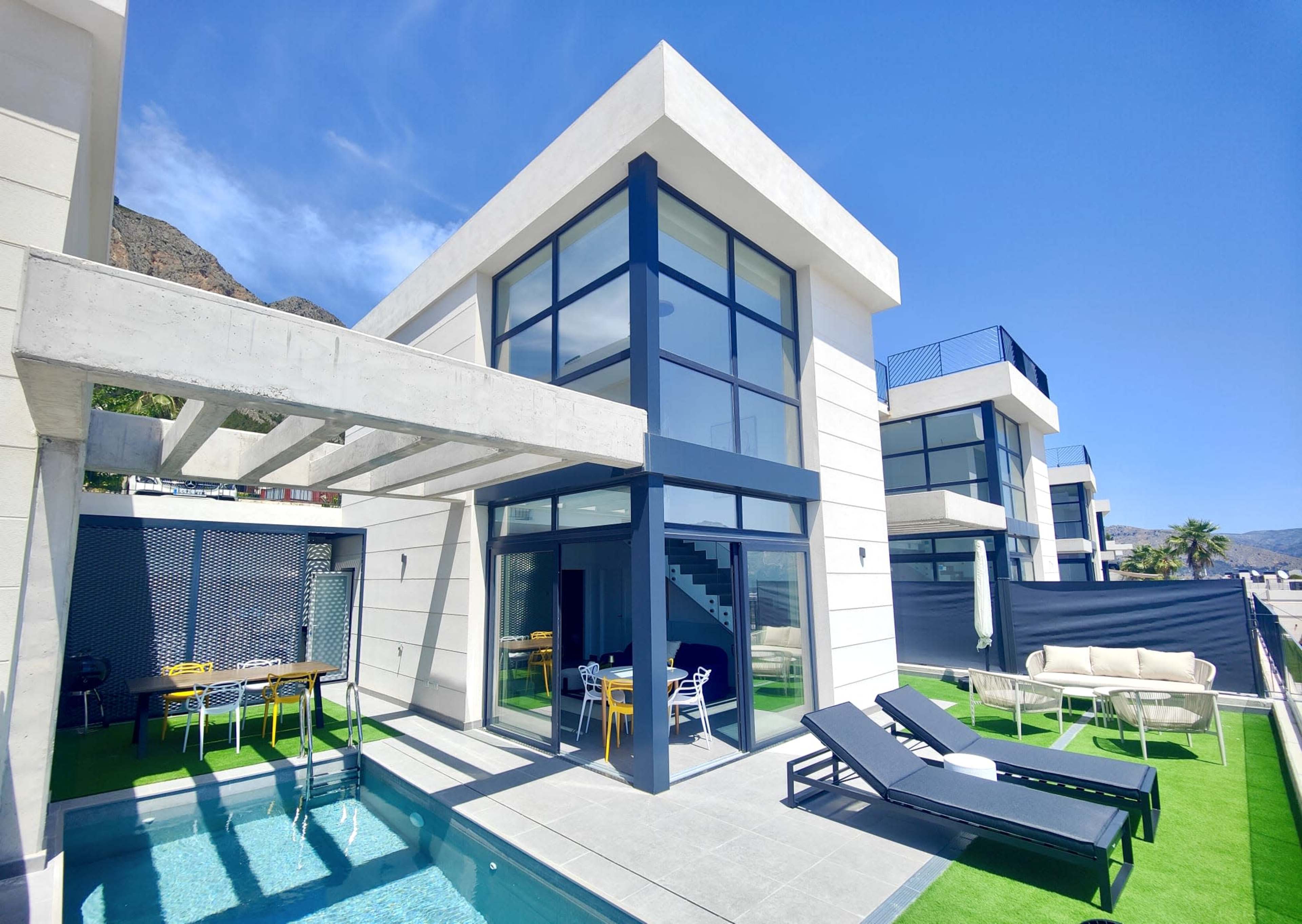 The image shows a modern two-story house with large glass windows, a small pool, and outdoor seating areas surrounded by a patio.