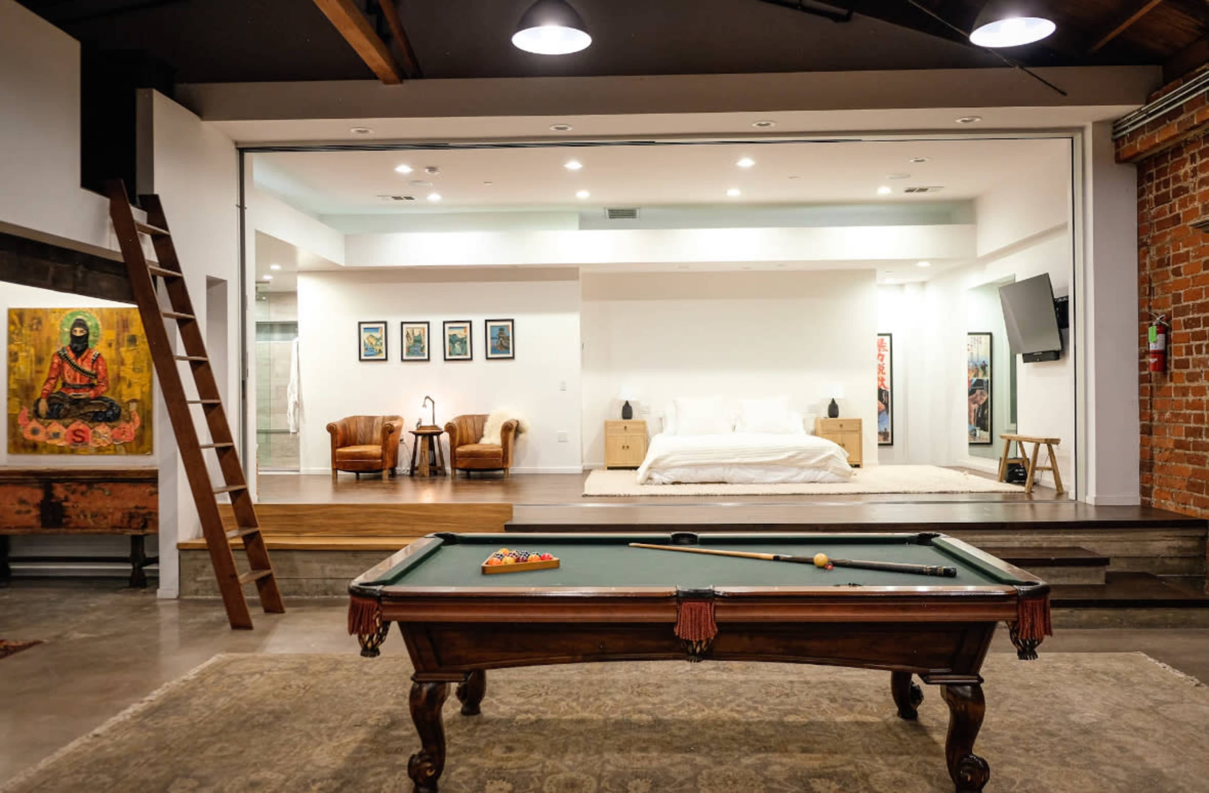 The image features a spacious loft with a pool table in the foreground and a minimalist bedroom area visible in the background.