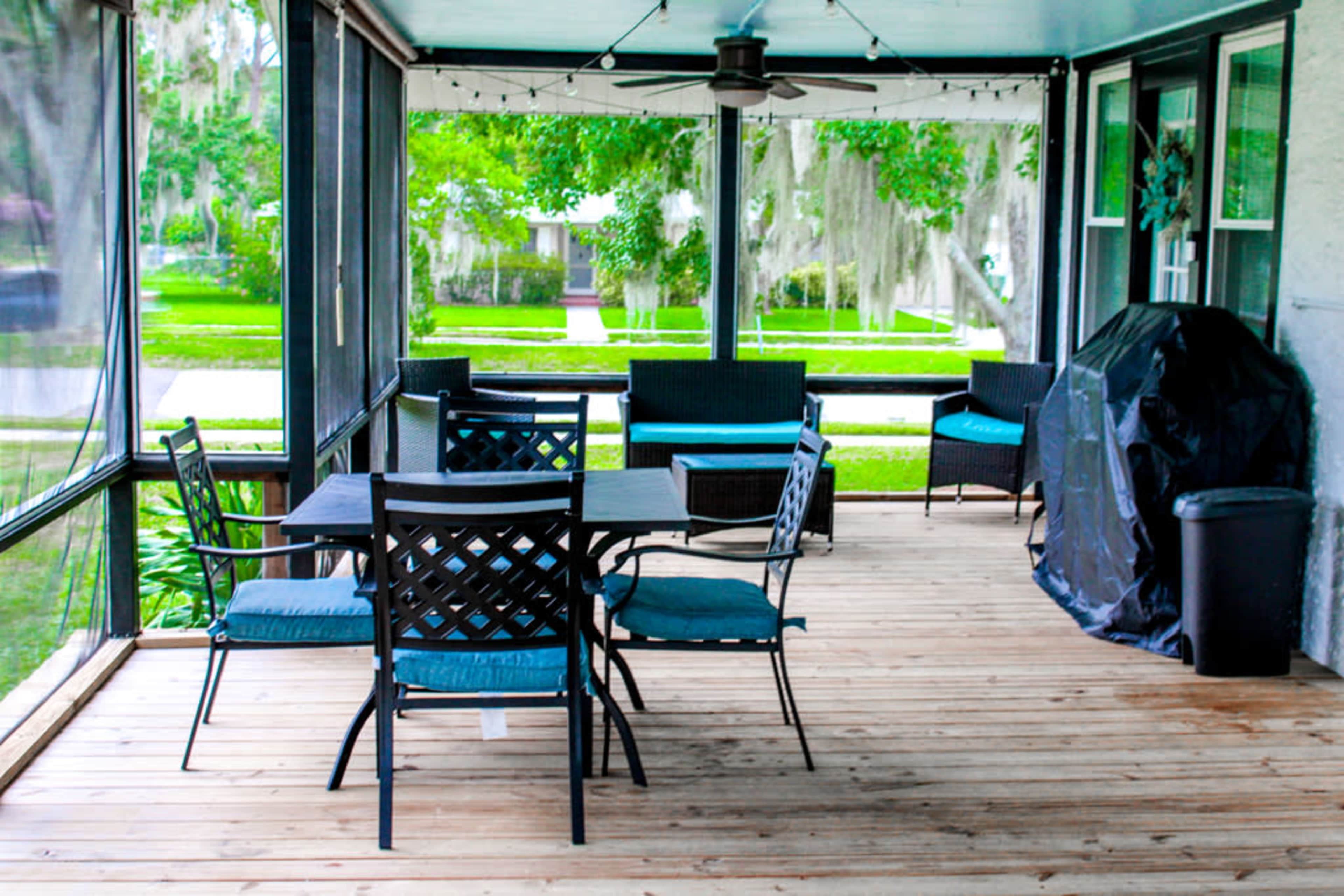 A screened porch featuring a wooden floor, a dining table with chairs, a seating area, and a charcoal grill.