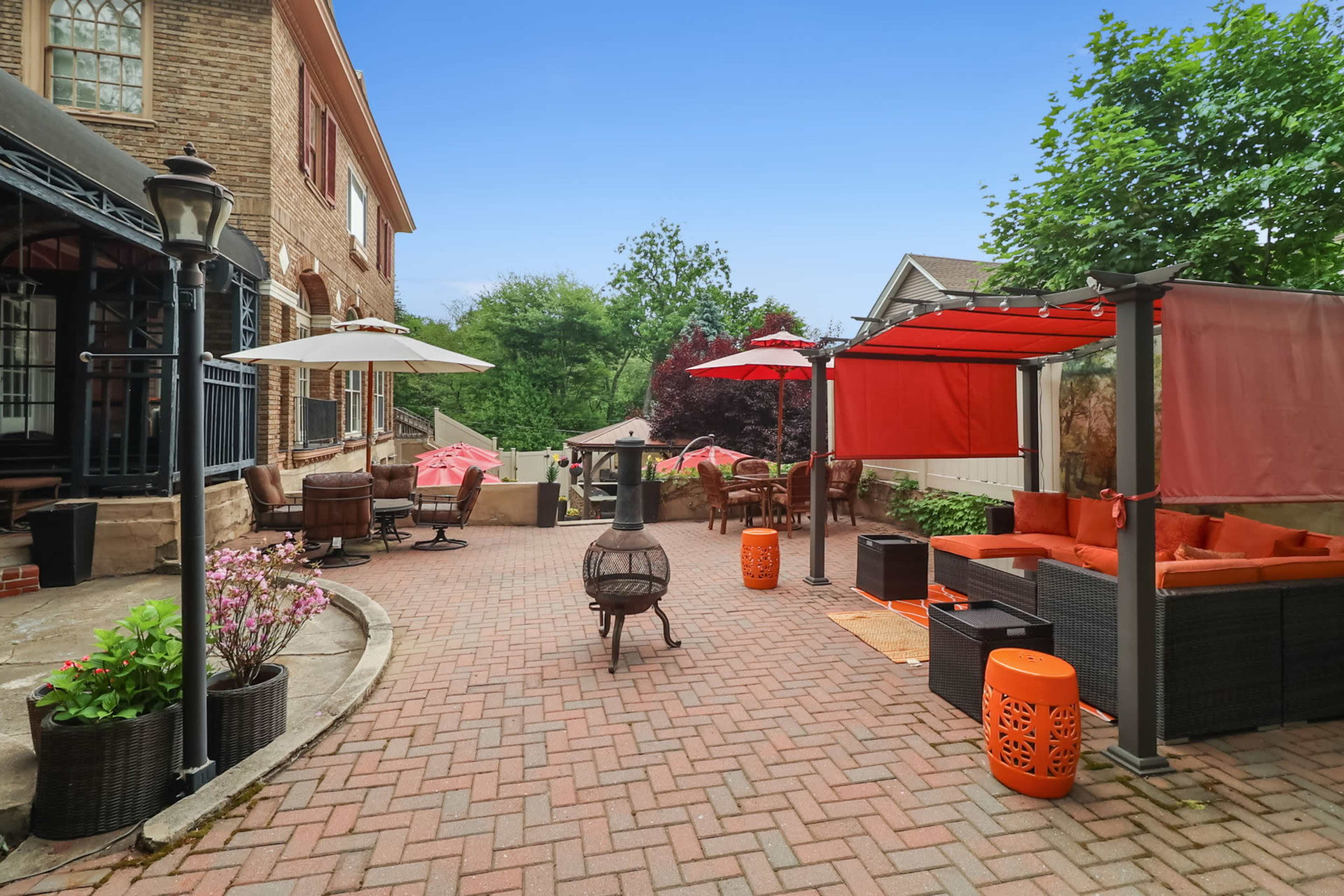 A brick patio features various seating areas with umbrellas and a fire pit, surrounded by greenery and decorative plants.