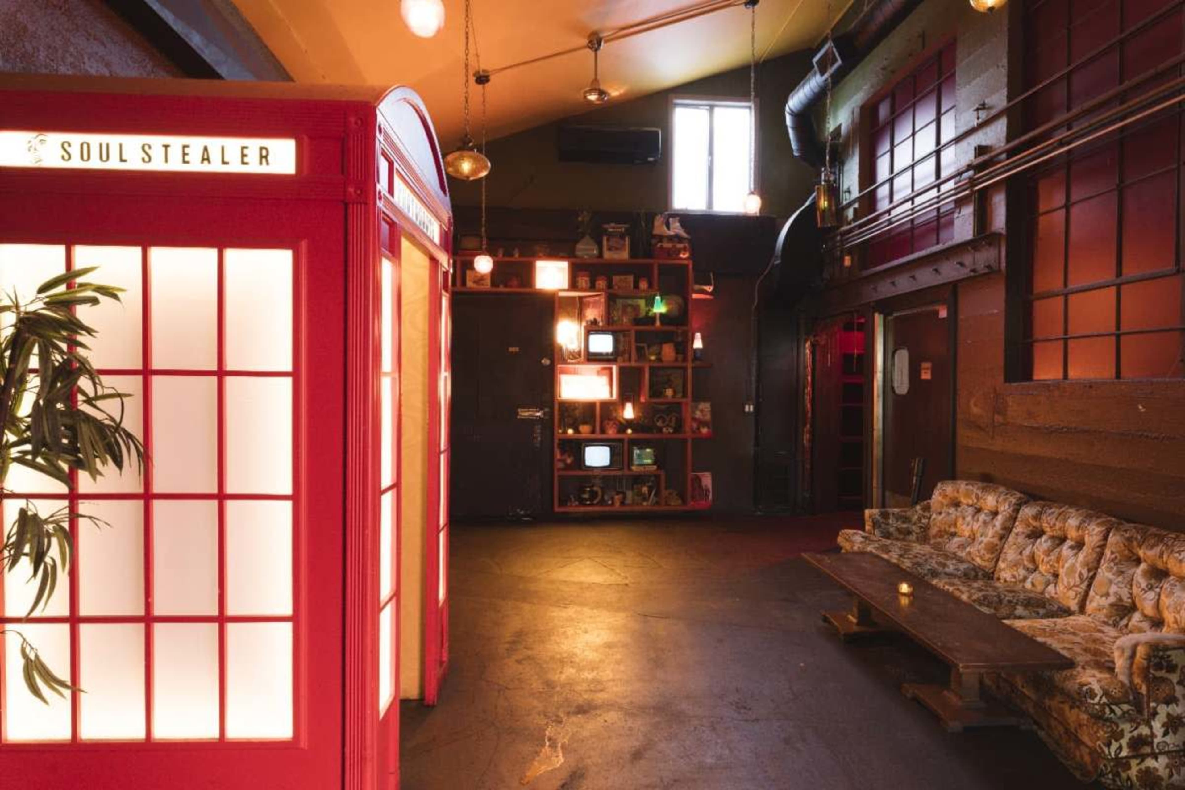 The image shows a vibrant, red telephone booth labeled "SOULSTEALER" inside a dimly lit, eclectic space with colorful shelves and a vintage sofa.