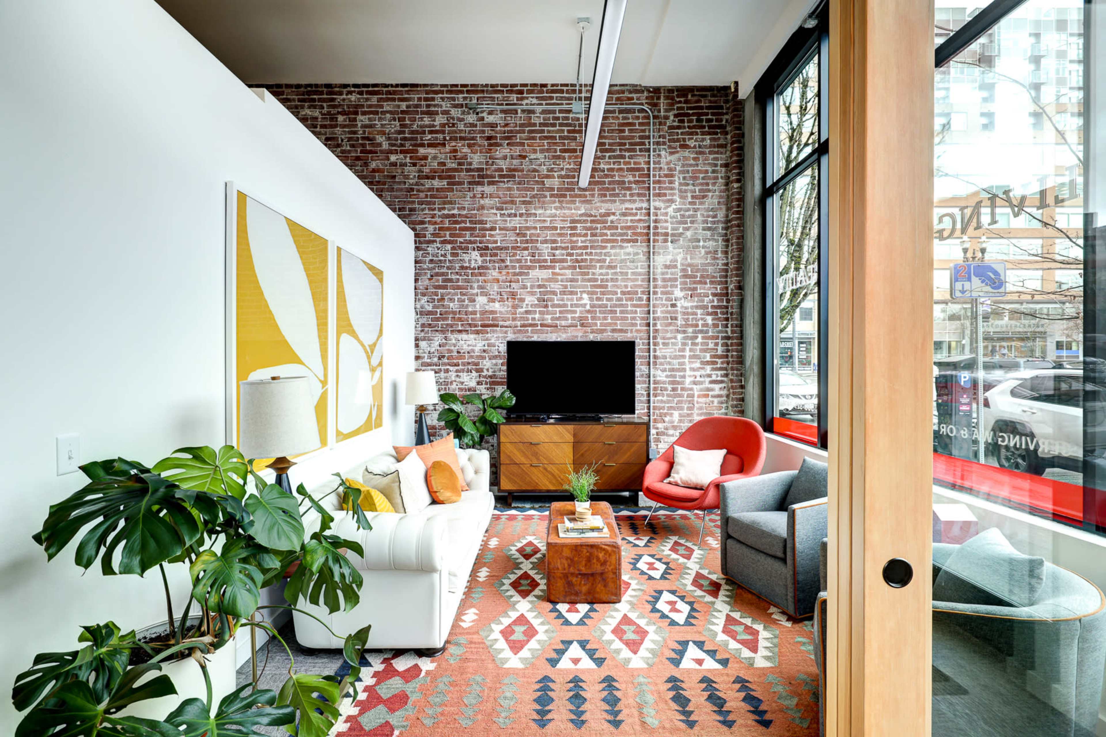 A bright living room features a brick wall, large windows, and modern furniture, including a white sofa, a red chair, and a wooden coffee table on a patterned rug.