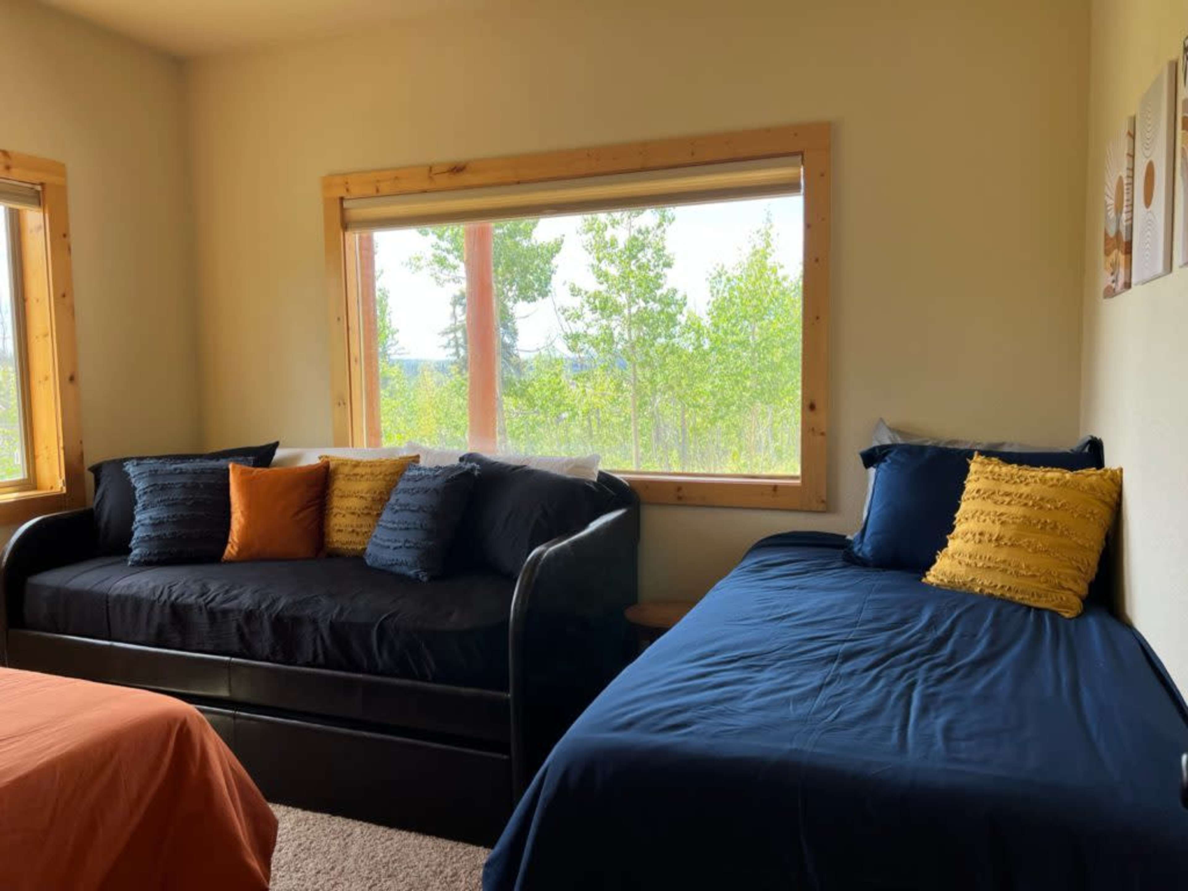 The image shows a cozy bedroom featuring two beds, one with a navy-blue cover and the other with black, both positioned near a large window that offers a view of trees outside.