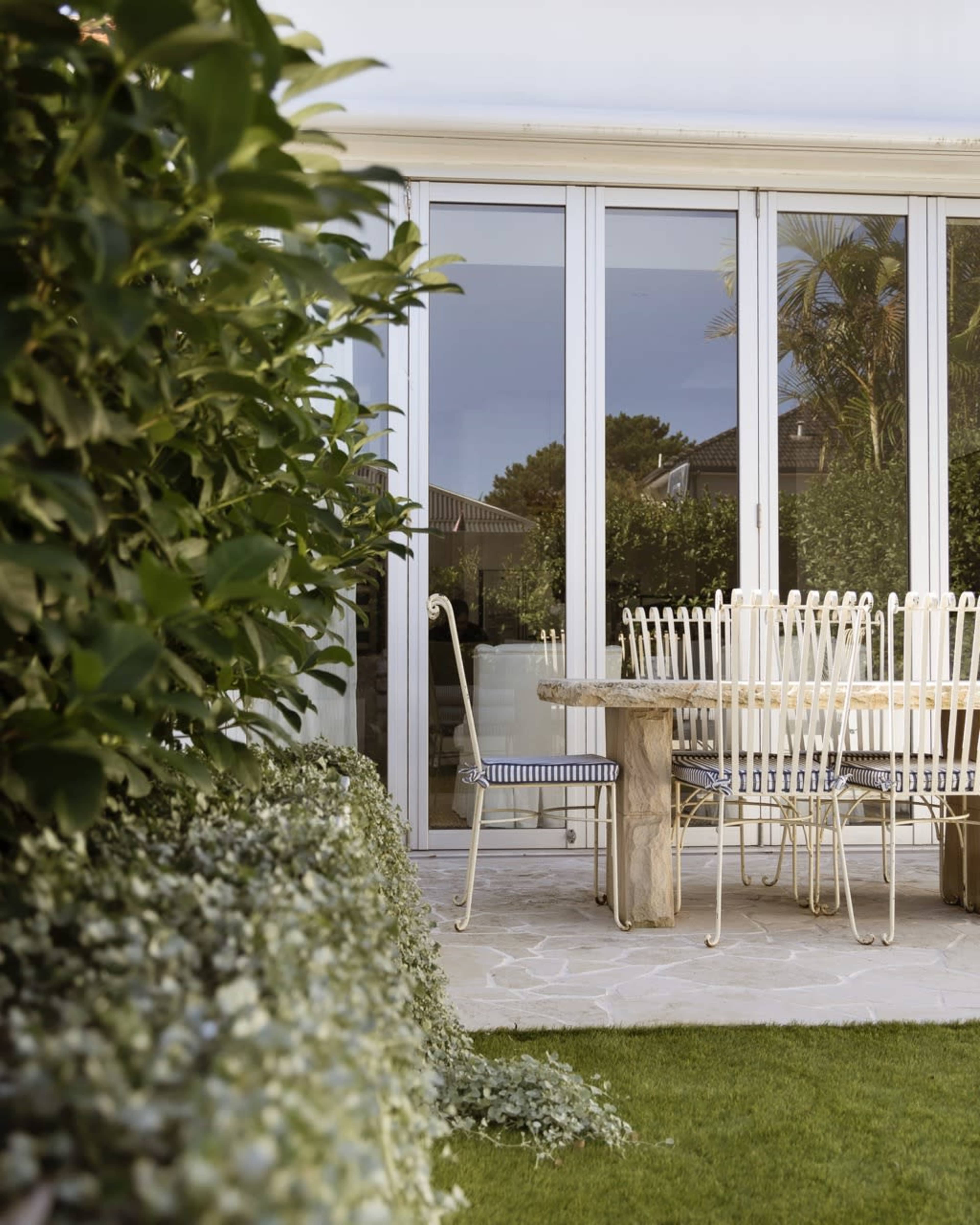 A patio features a wooden table surrounded by white metal chairs, bordered by lush greenery and large glass doors.