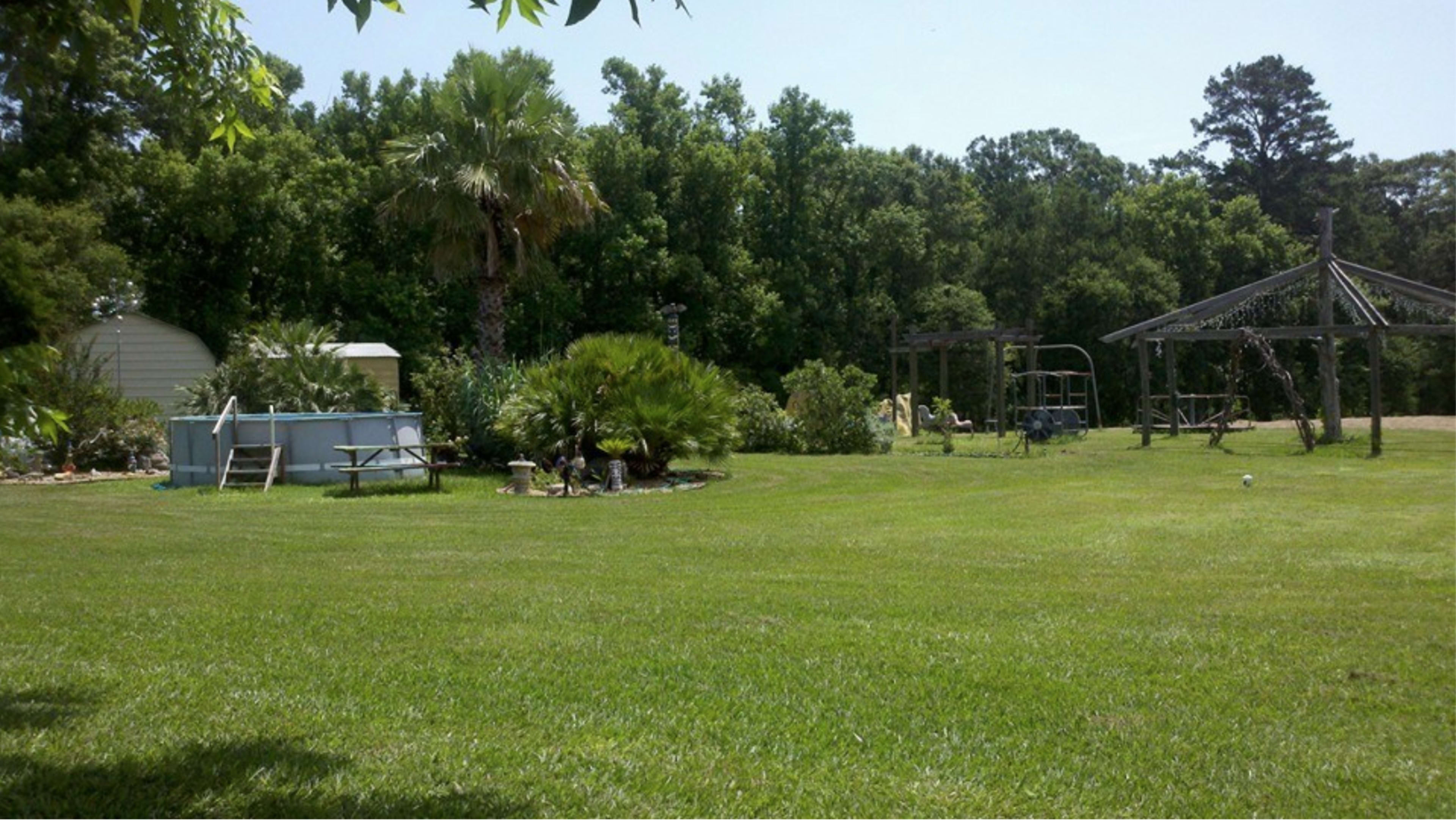 A well-manicured grassy yard featuring trees, a swimming pool, garden beds, and a wooden gazebo structure.