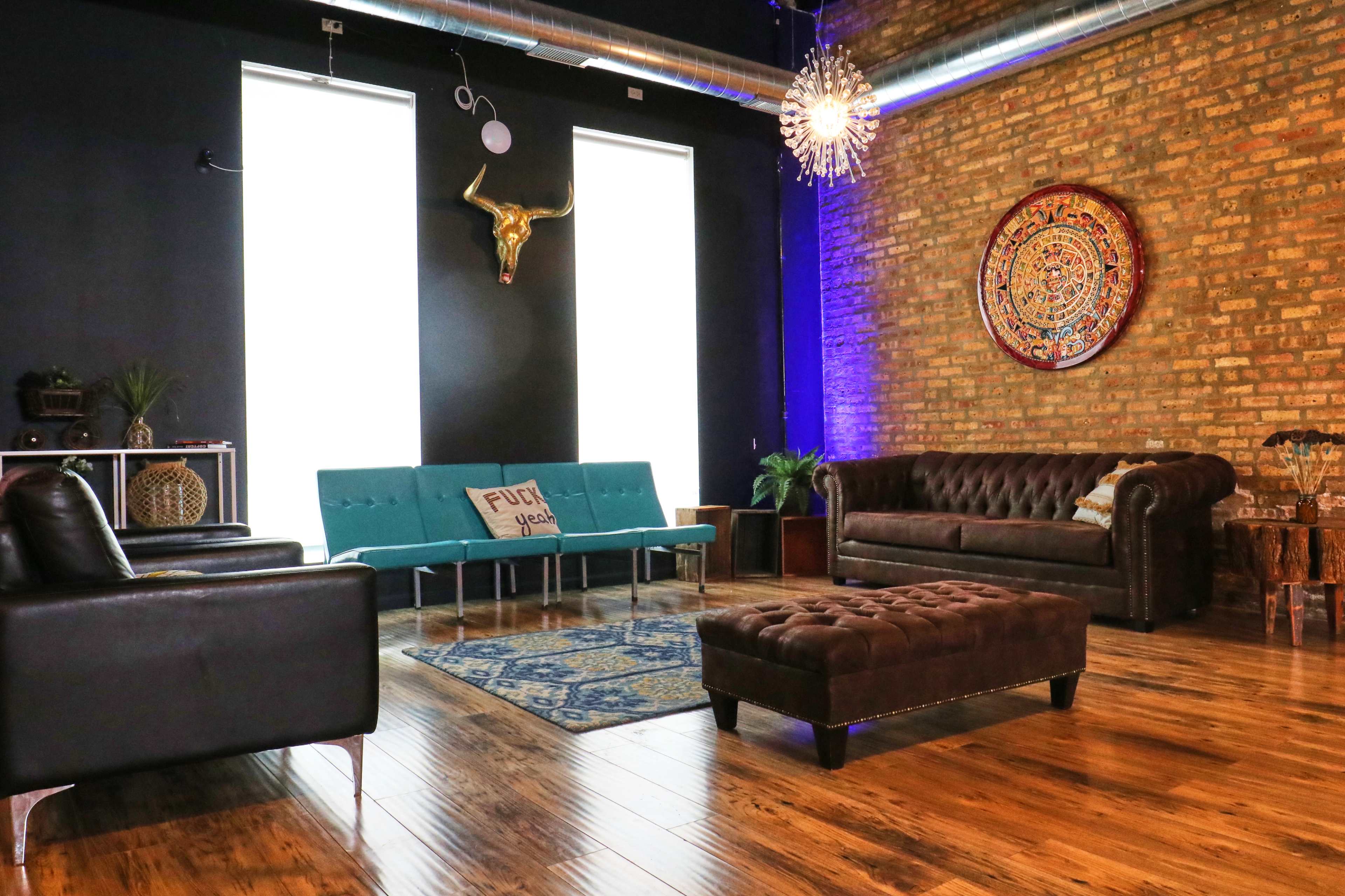 The room features a mix of modern and vintage furniture, including a blue sofa, a brown leather couch, and a tufted ottoman, against a backdrop of exposed brick walls and large windows.