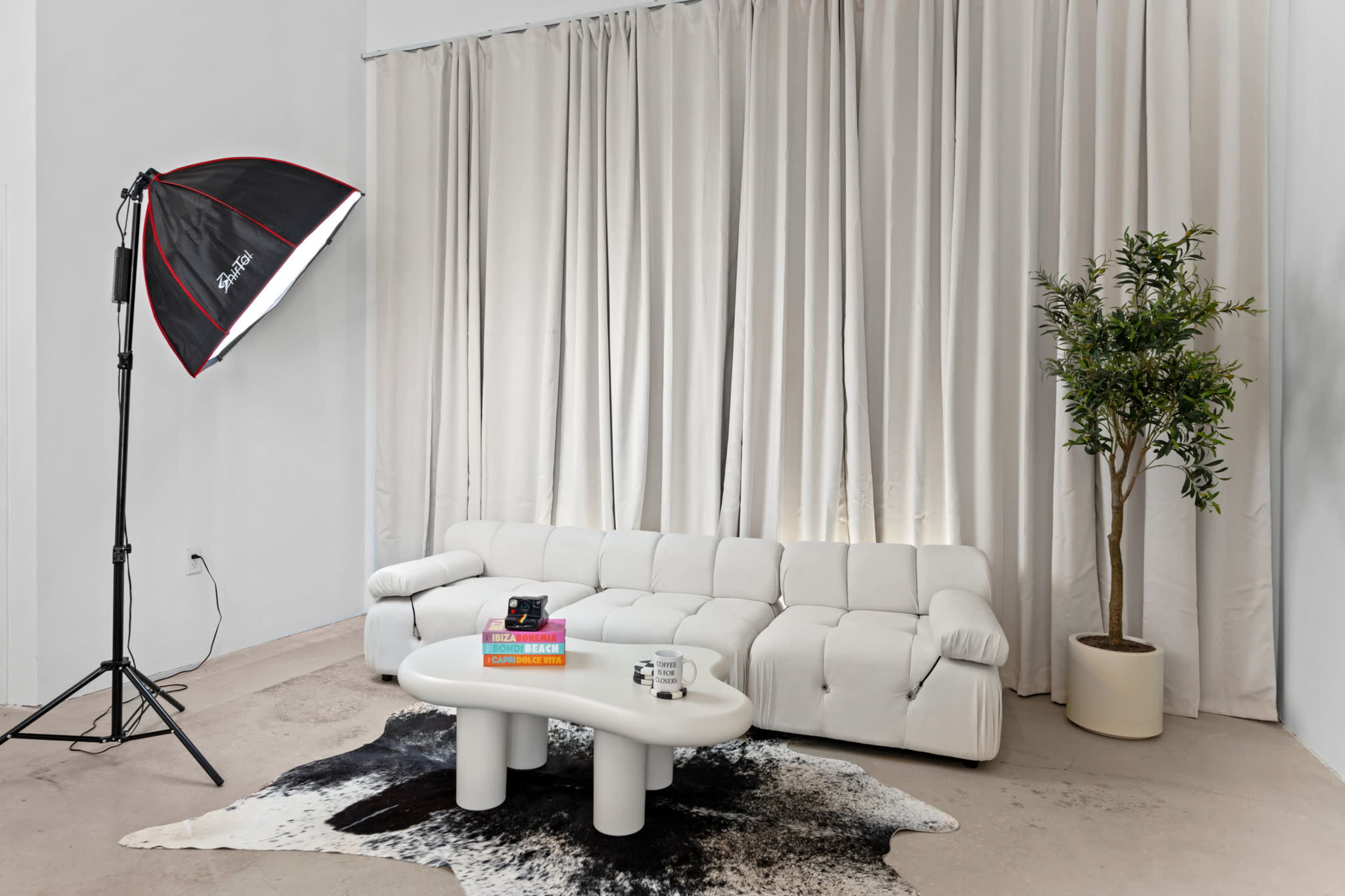 The scene features a white sectional sofa with a round coffee table, a potted plant, and a large softbox light directing towards the corners, all set against a backdrop of white curtains.