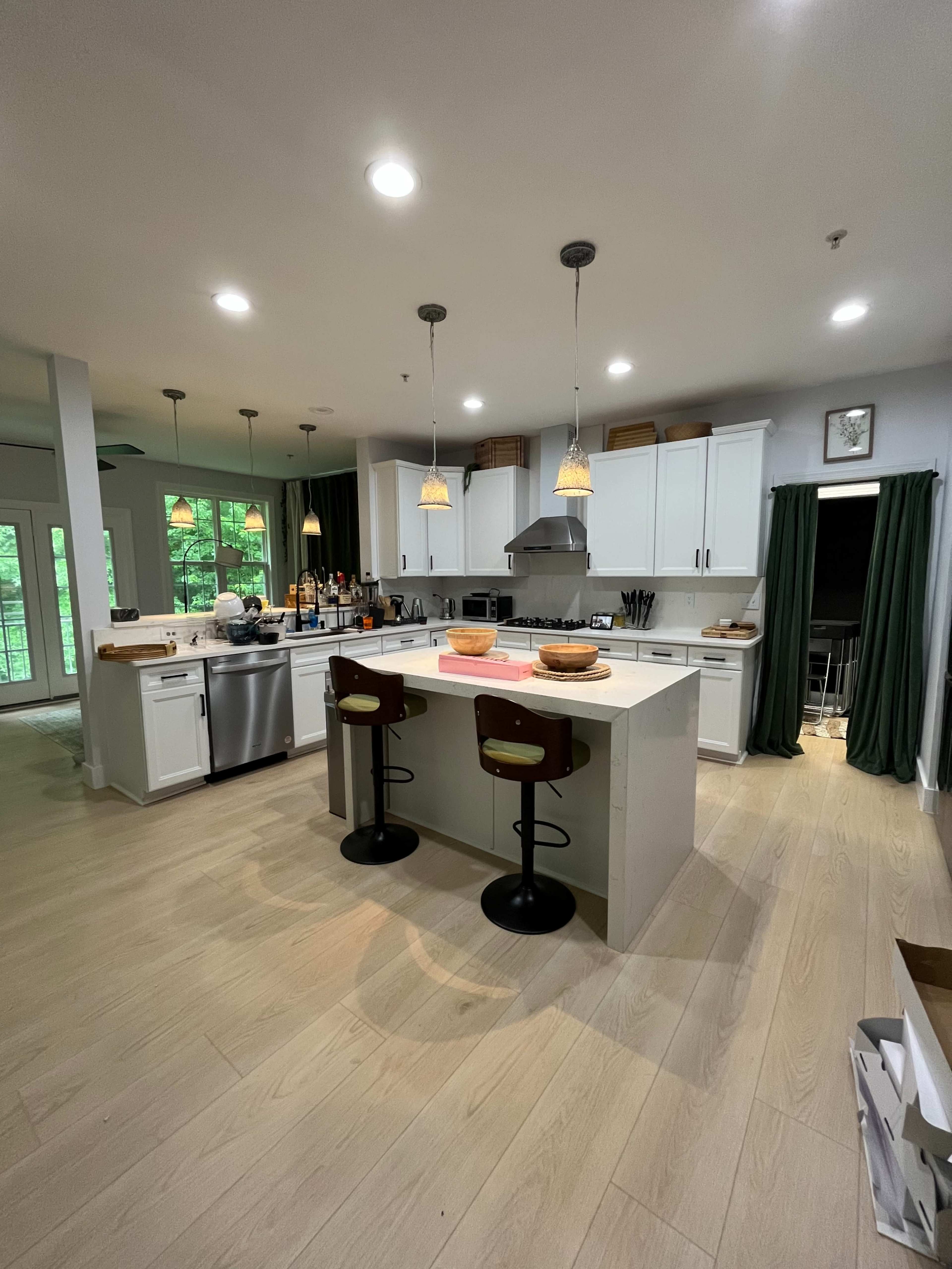 The image shows a modern kitchen with a central island, two bar stools, white cabinetry, and large windows allowing natural light.