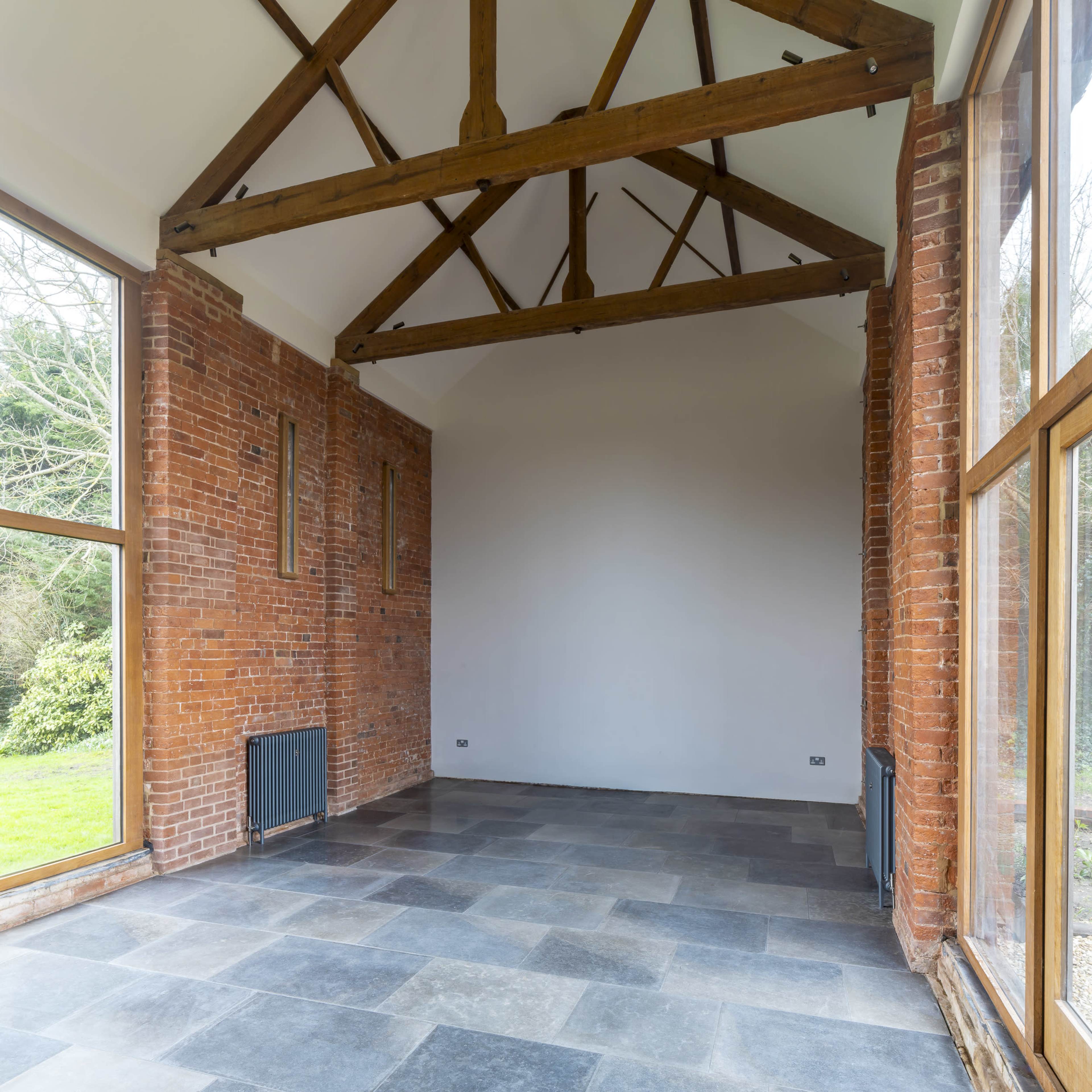 The image shows a spacious room with high wooden beams, brick walls, and large windows, featuring a stone floor.