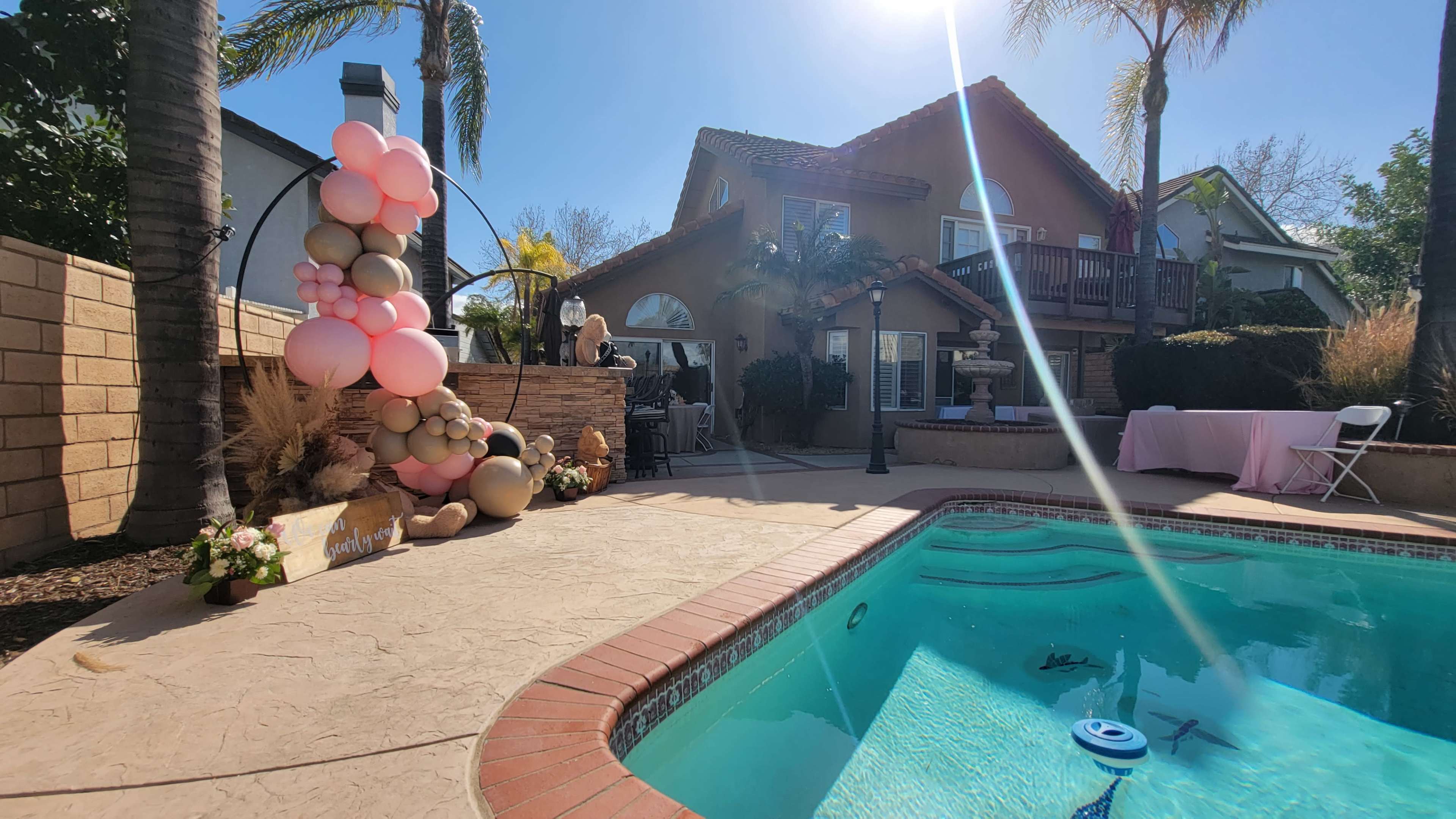A poolside scene features a decorated area with a balloon arch, a patio with seating, and a house in the background under clear blue skies.