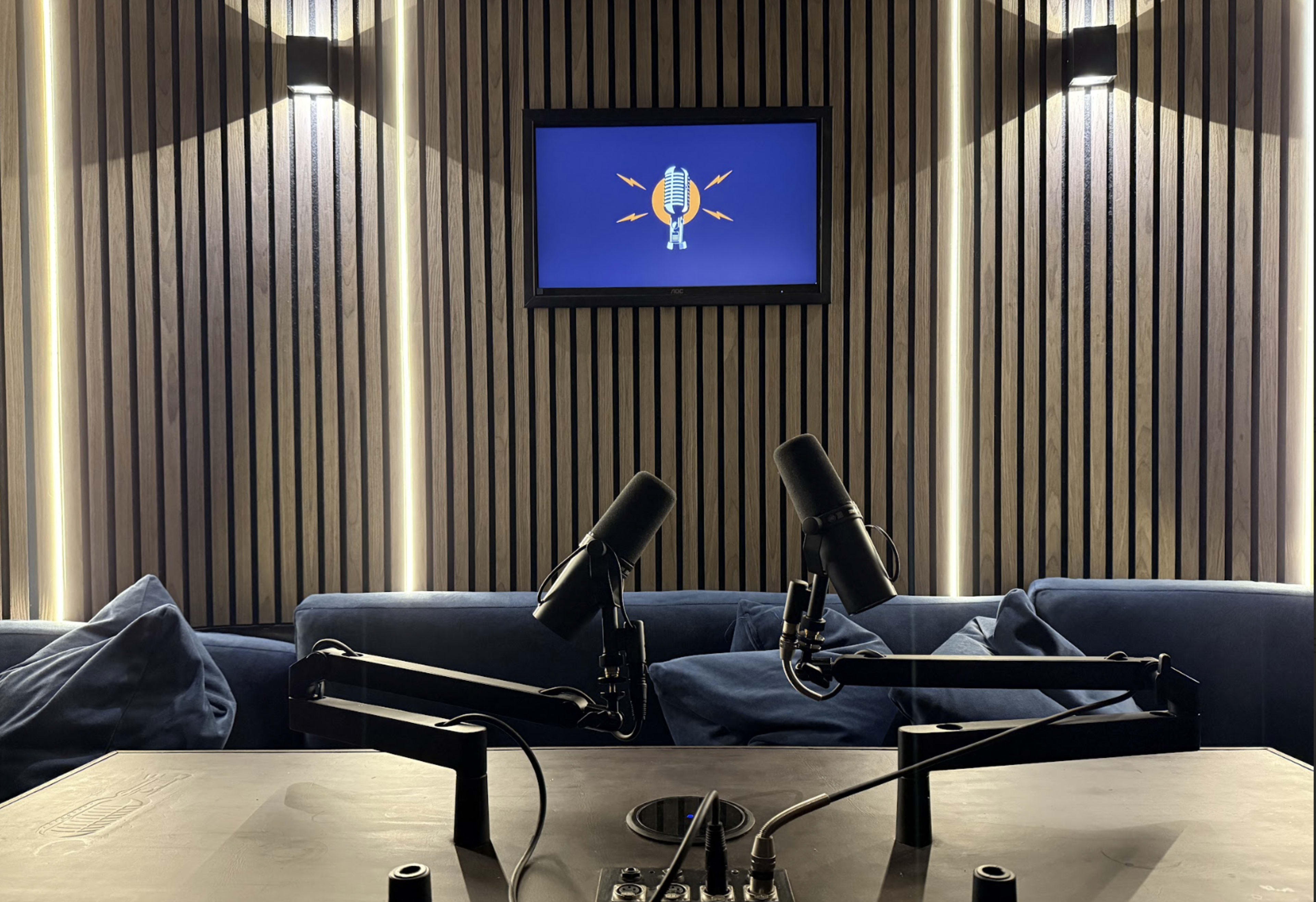 The image shows a podcast recording studio with two microphones mounted on adjustable arms and a screen displaying a graphic in the background.