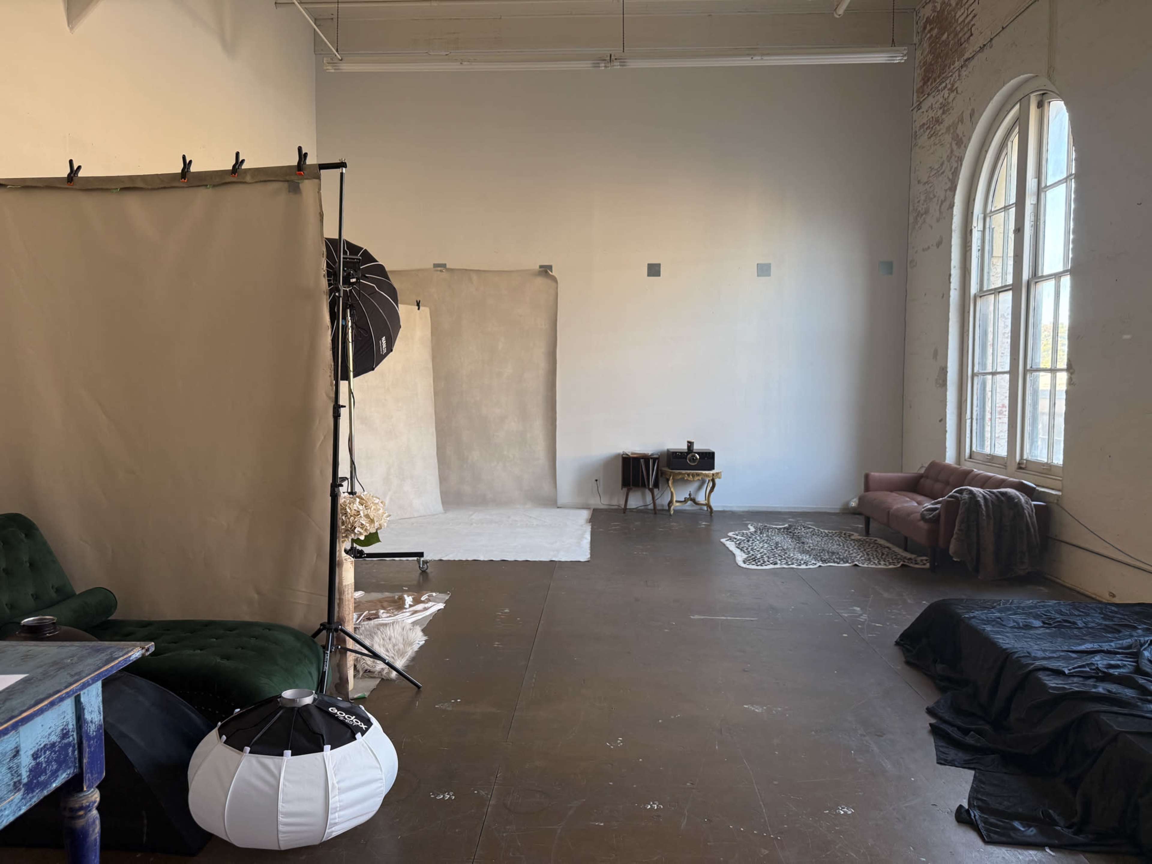 The image shows a photography studio with a backdrop setup, soft lighting equipment, and a few seating options in a spacious, well-lit room.