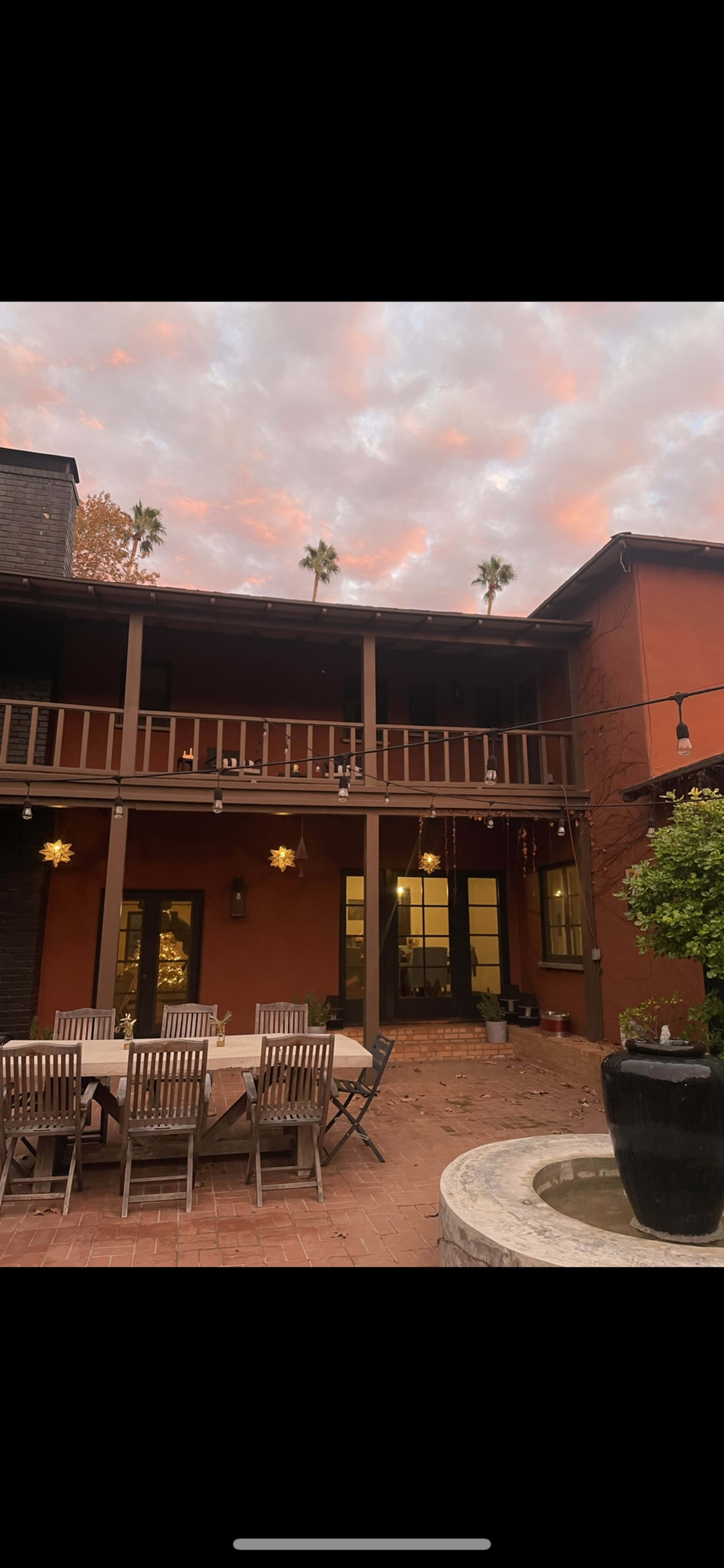 A two-story building with orange walls, featuring a balcony and a patio set with wooden chairs and a table, surrounded by palm trees and a fountain.