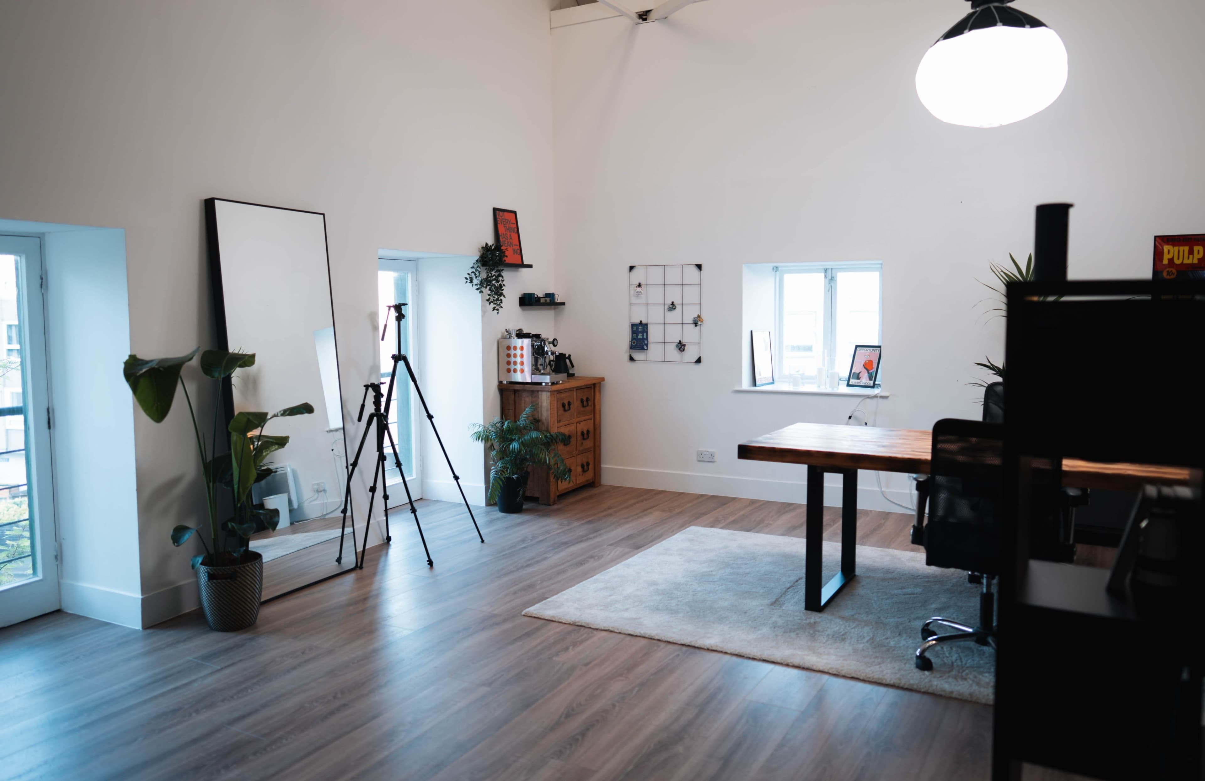 A bright, spacious office features a large wooden desk, a mirror, a tripod, and several plants against white walls and wooden flooring.