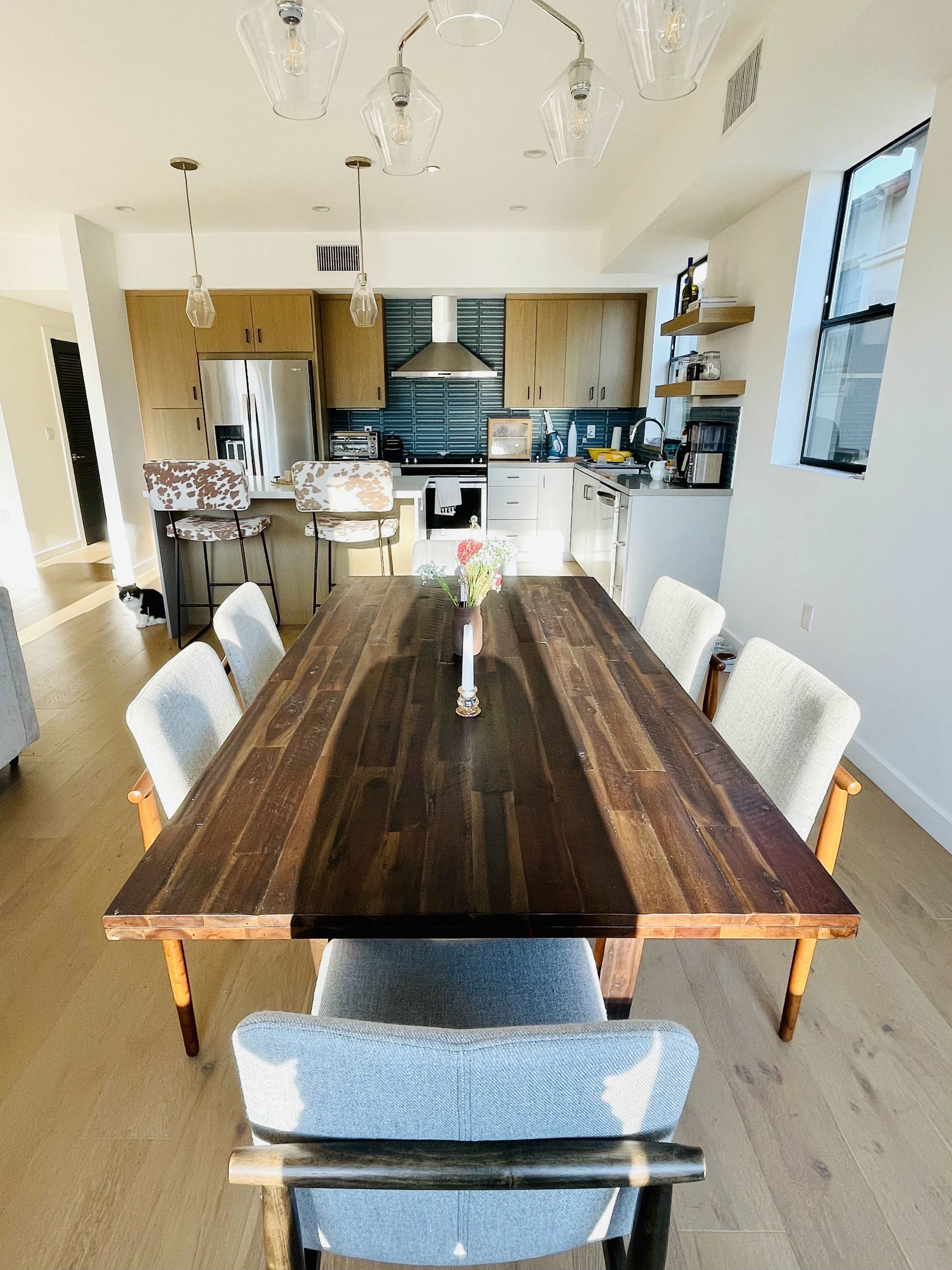 A wooden dining table is set in a modern kitchen with open shelving, light fixtures, and a contrasting color scheme.