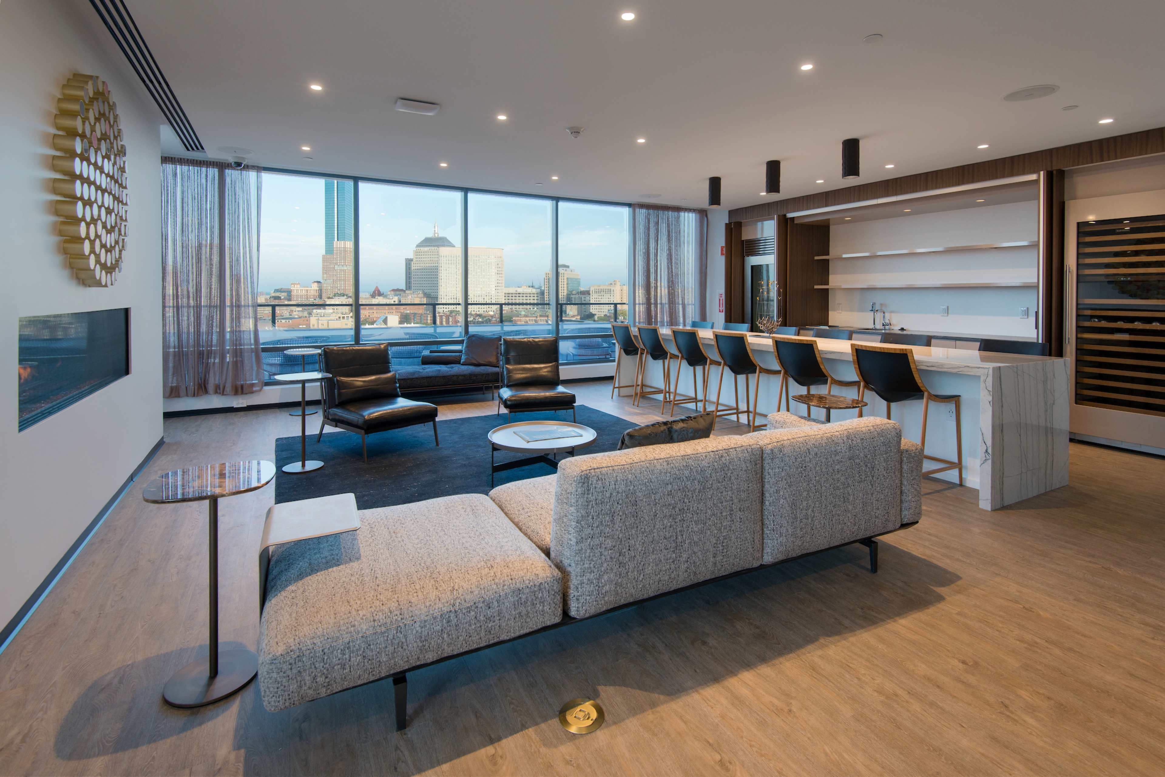 A modern living space features a sectional sofa, a glass table, and a bar area with high stools, along with a view of a city skyline through large windows.