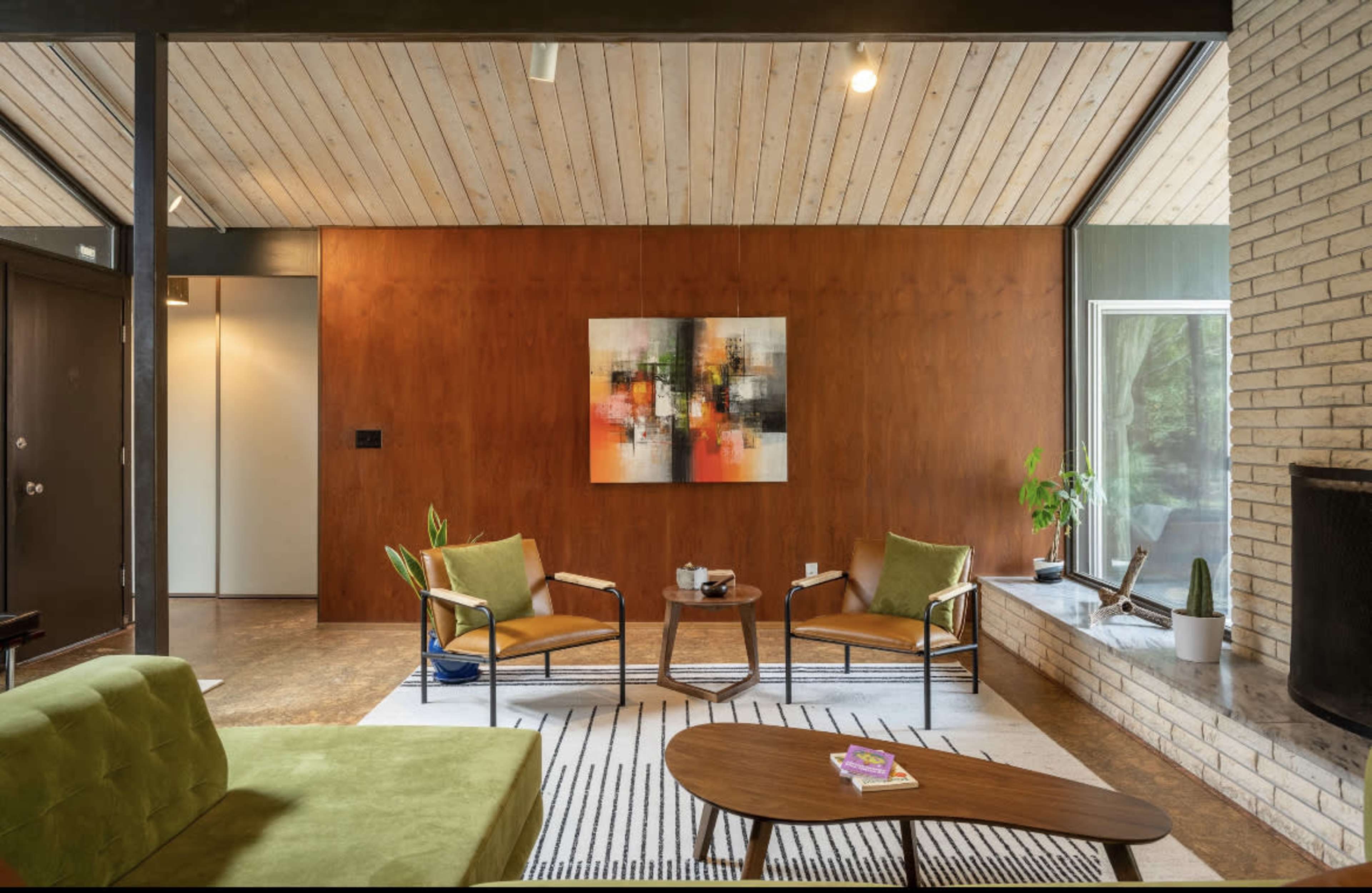 A modern living room featuring two green chairs and a sofa, a wooden coffee table, and a large artwork on a brown wall.