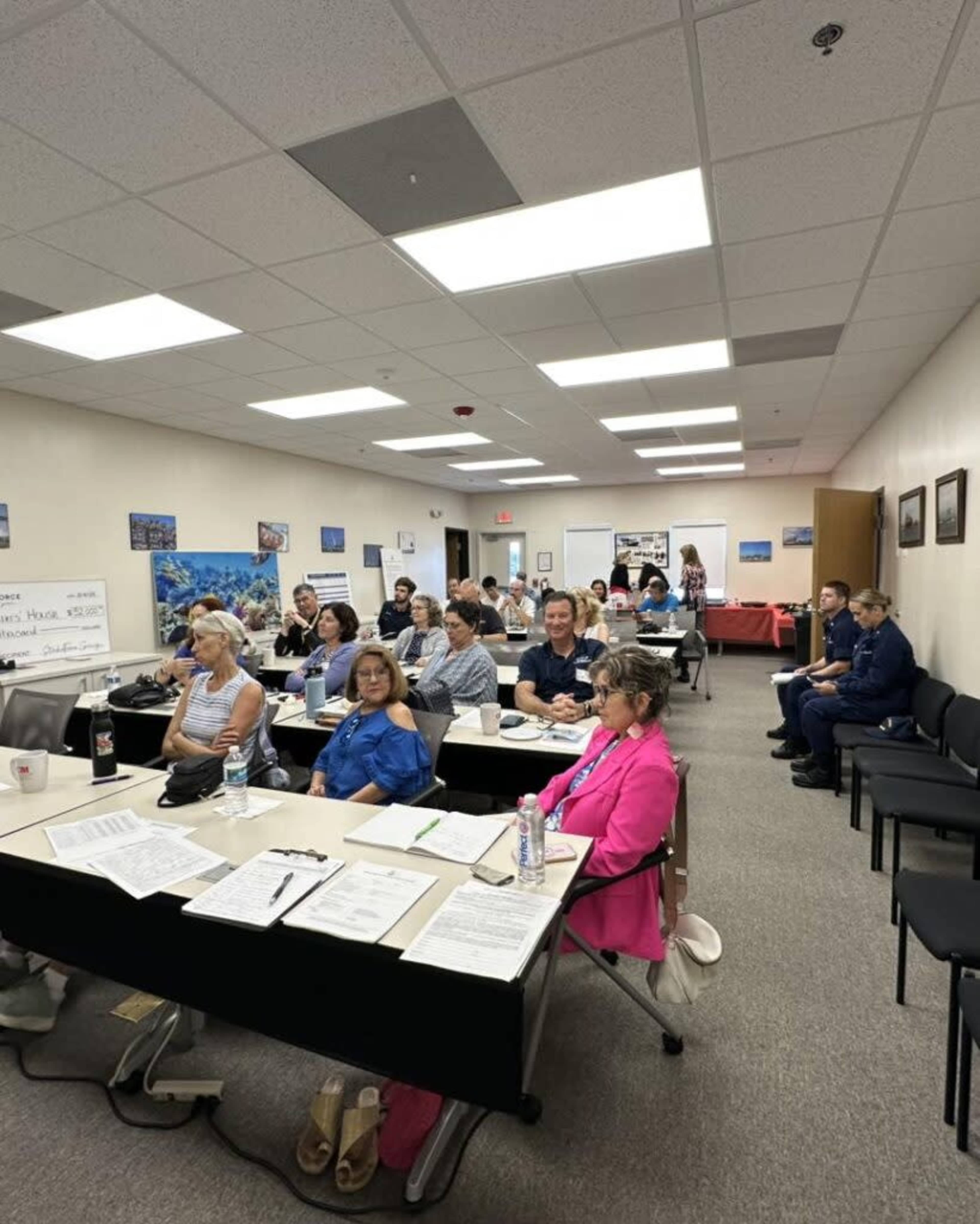 A group of people sits in rows of tables in a classroom setting, attentively listening to a speaker at the front of the room.