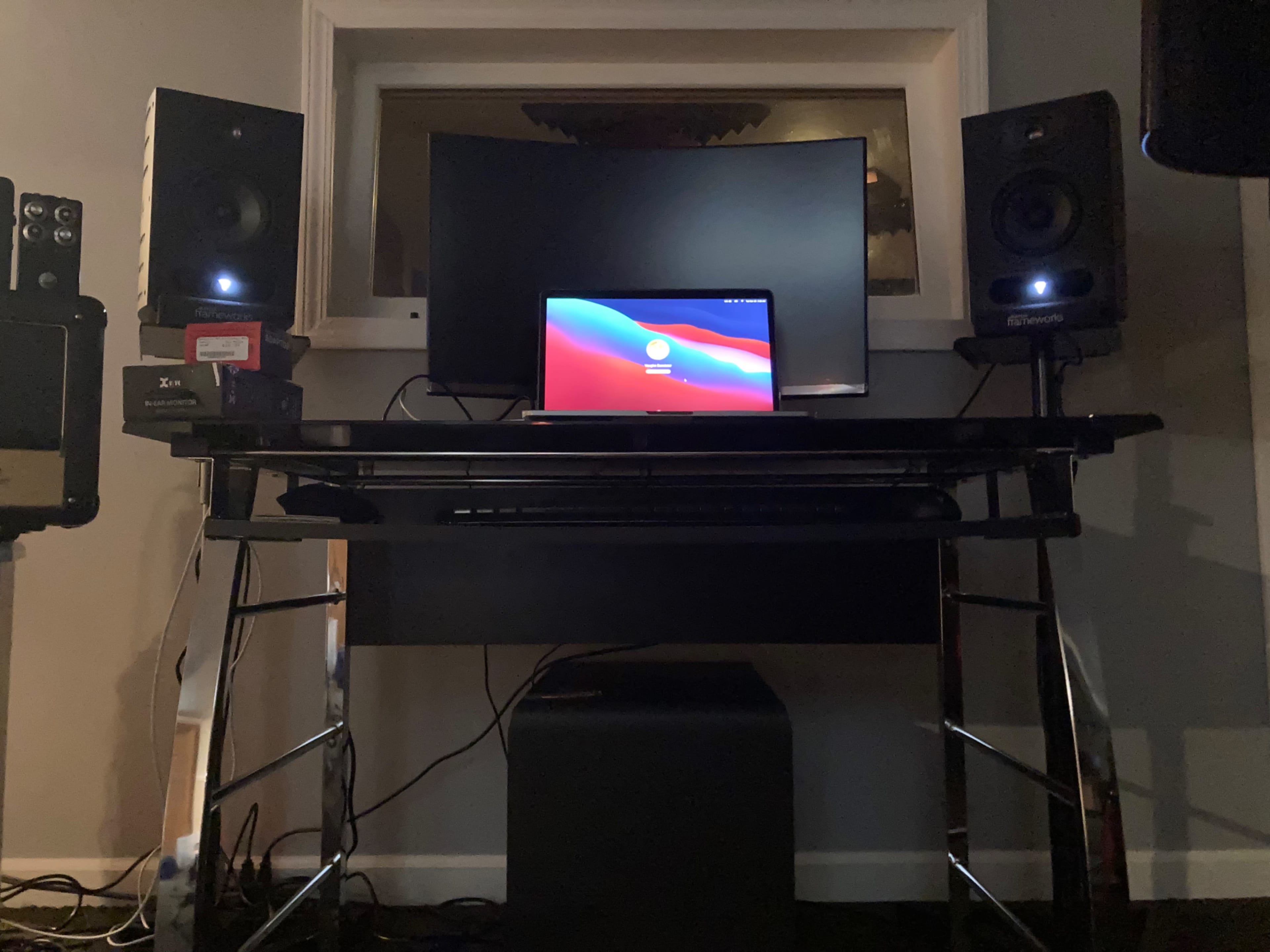 A desktop setup features a curved monitor, laptop, and two speakers on a black table with visible cables.